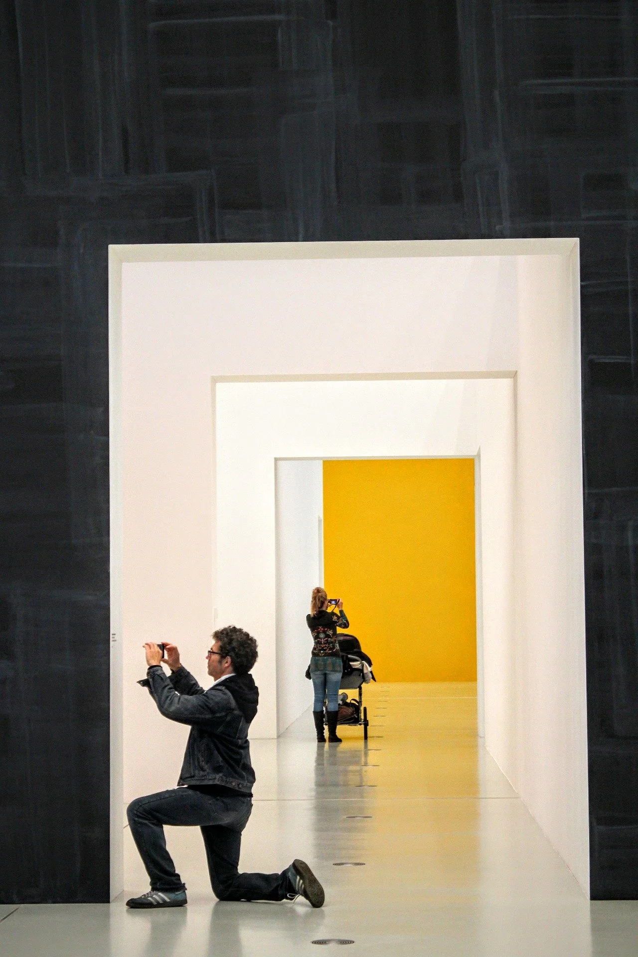 People taking photos in an art gallery, with one person kneeling and another standing in front of a yellow art installation framed by a white structure.
