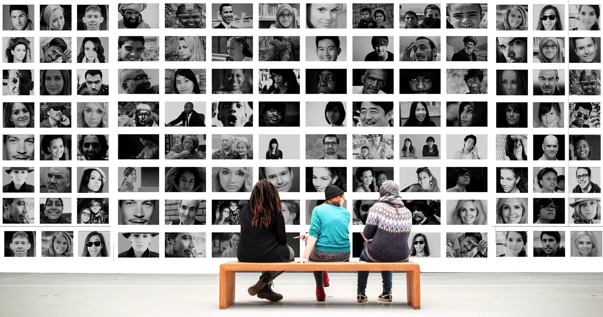 Three people sitting on a bench in front of a large wall collage of black and white portrait photographs of diverse people.