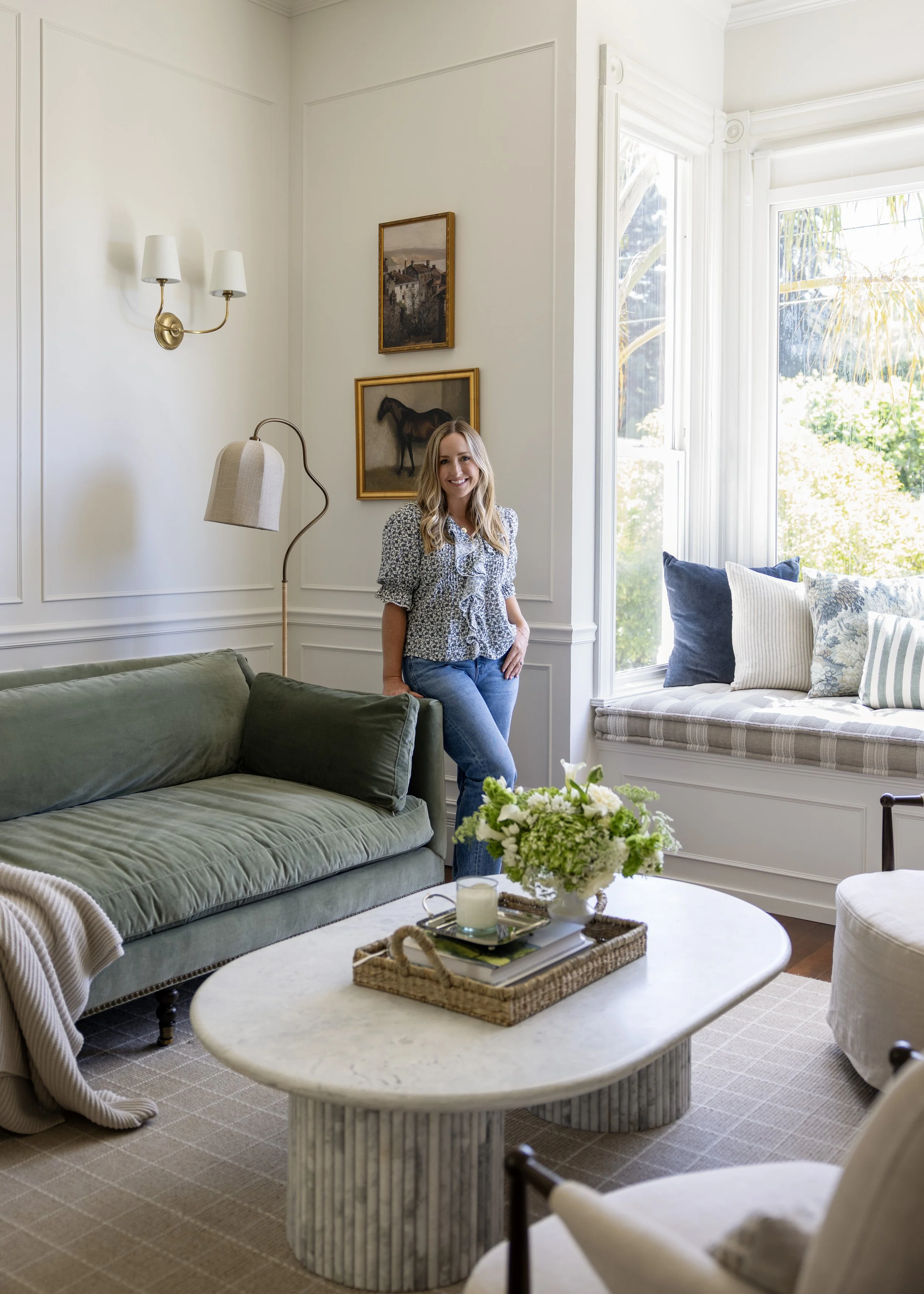 A woman with blonde hair, wearing a patterned blouse and jeans, standing in a bright living room next to a large window with white trim. The room has white walls with decorative molding, artwork featuring a house and a horse, a beige floor lamp, a green sofa, a glass coffee table with a bouquet of white and green flowers, and a beige patterned rug.