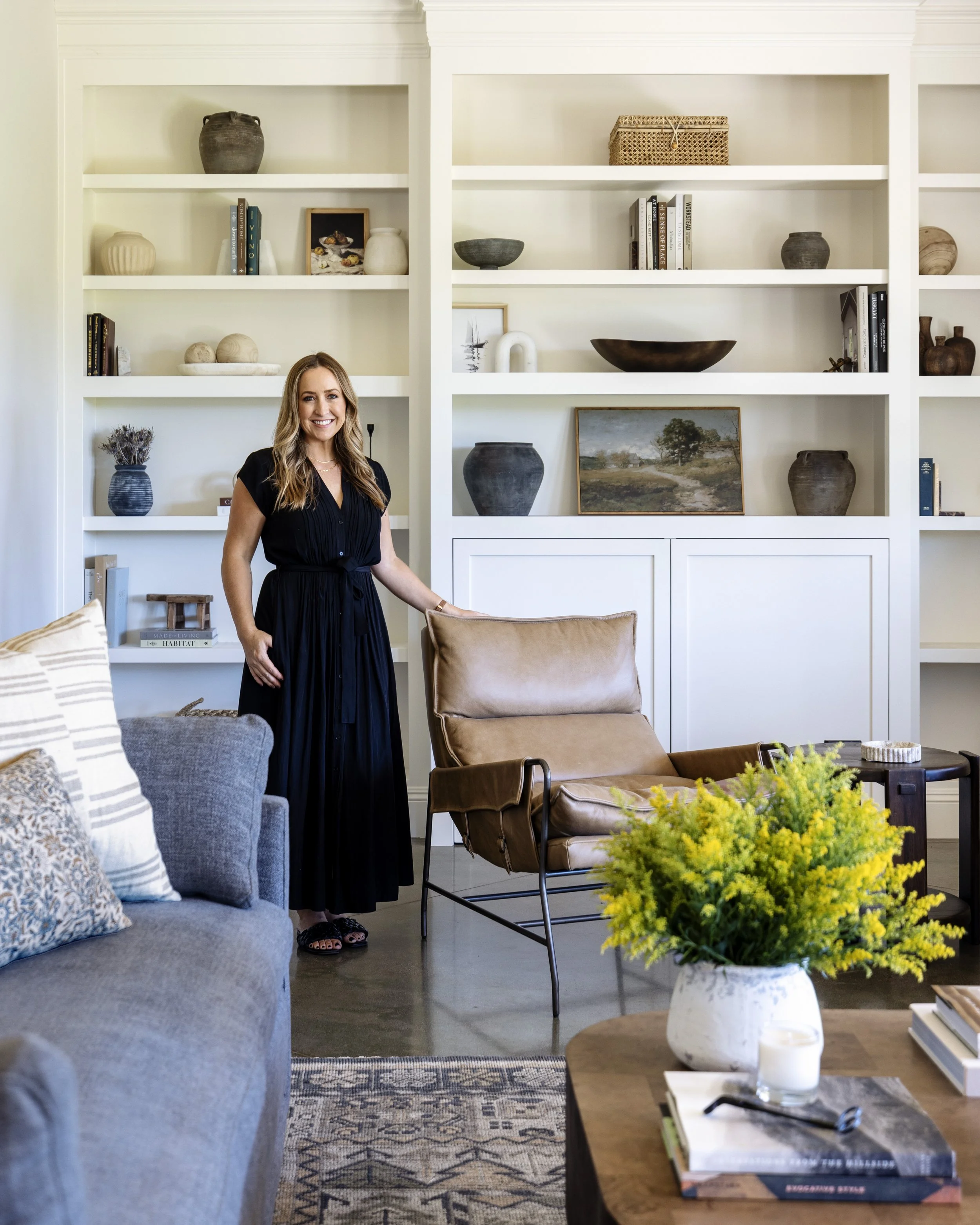 A woman with long blonde hair smiling in a black dress standing in a modern living room. The room has bookshelves with decorative items, a gray sofa with patterned pillows, a wooden coffee table with yellow flowers in a white vase, and a stack of books with a black pair of glasses on top.