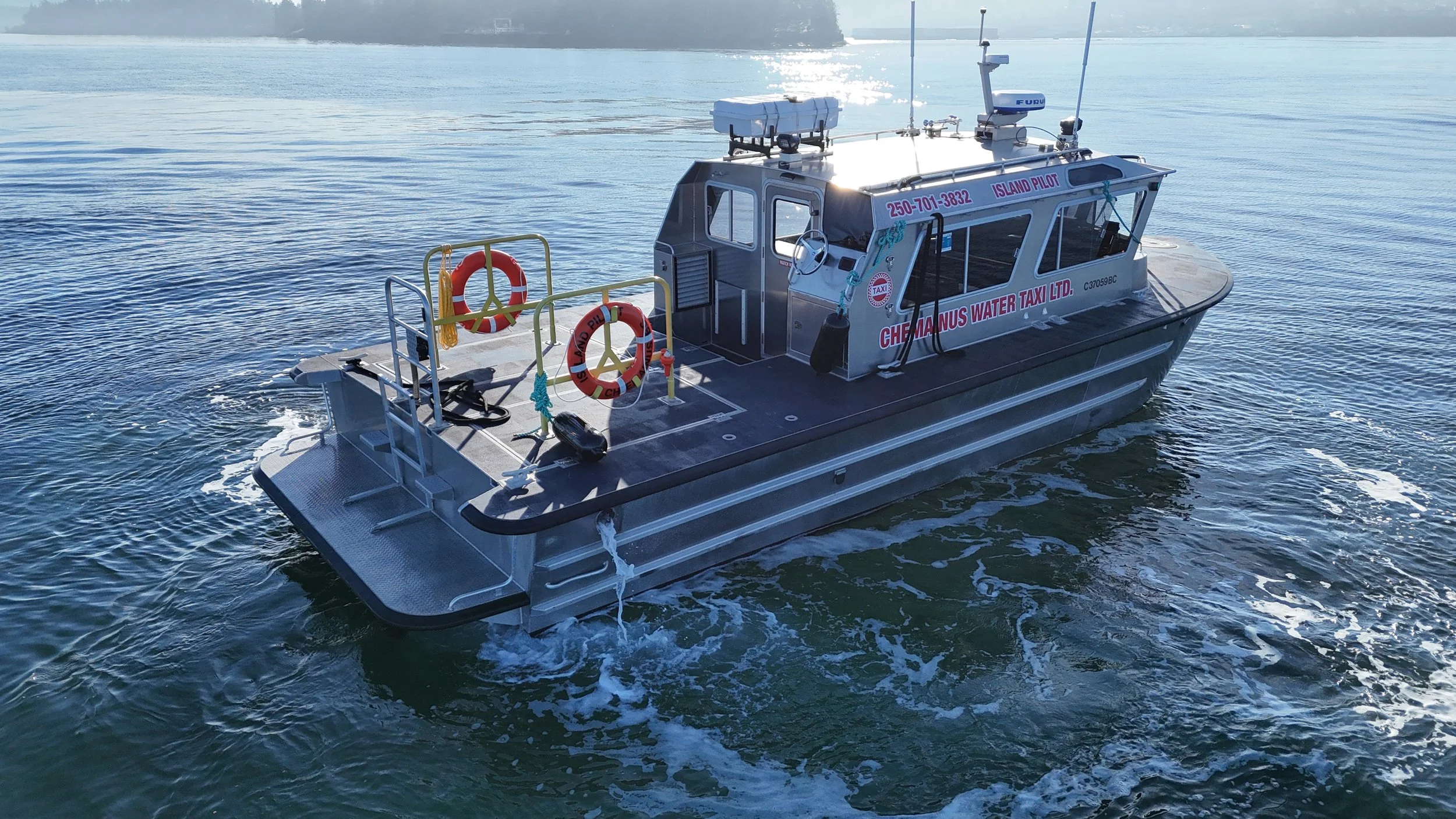 A modern water taxi boat named Chemainus Water Taxi LTD is on the water, with a visible phone number 250-701-3832. The boat is equipped with safety rings, antennas, and electronic equipment, with a scenic background of water and distant shoreline.