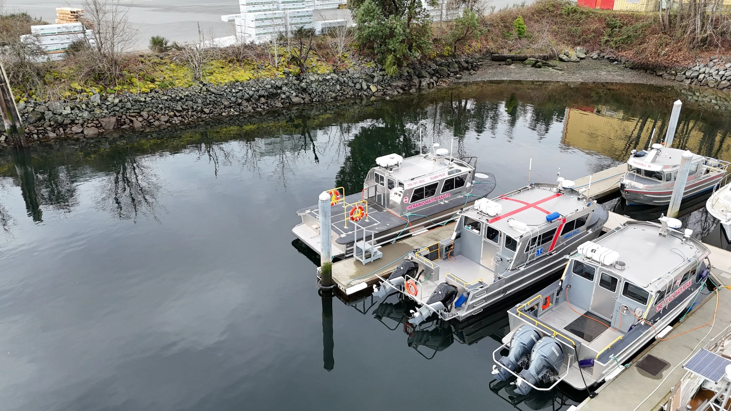 Several boats docked at a marina, with an island pilot and ambulance boat among them. The water is calm, reflecting the boats and nearby trees and structures.