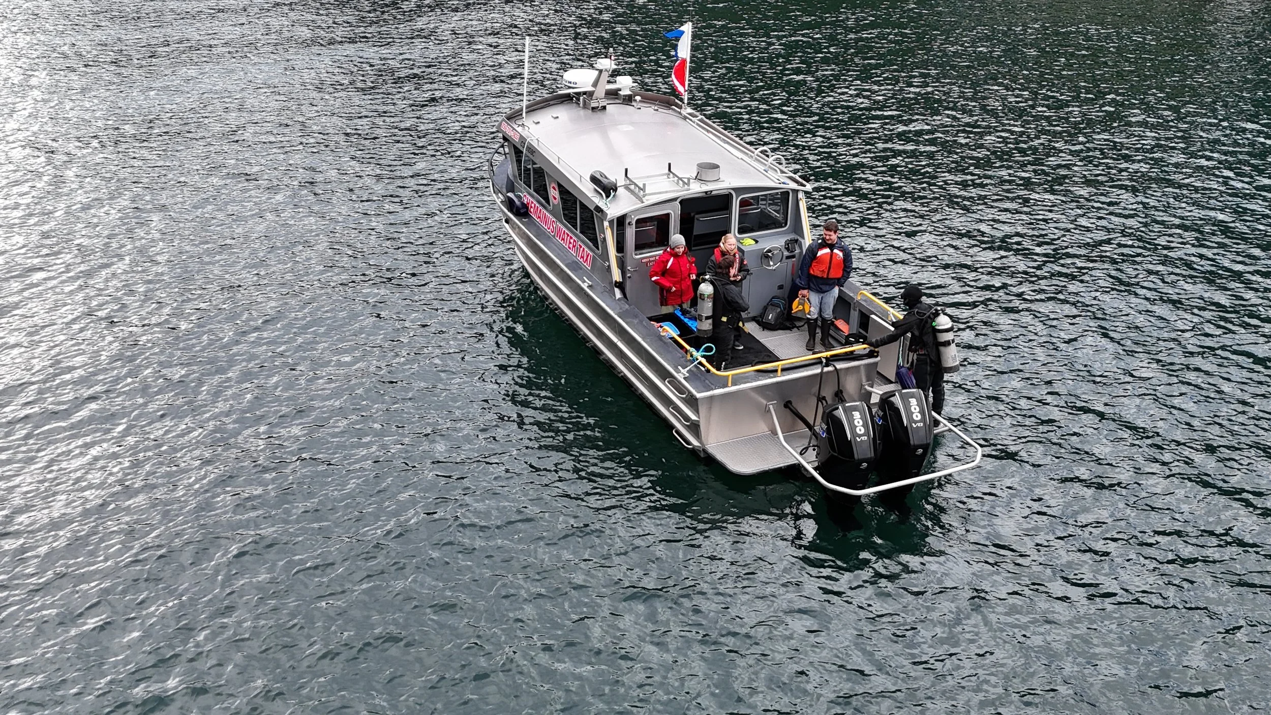 A water taxi with four passengers and crew members on board, floating on a body of water. The boat is labeled 'Vienna Water Taxi' and is equipped with two outboard motors.