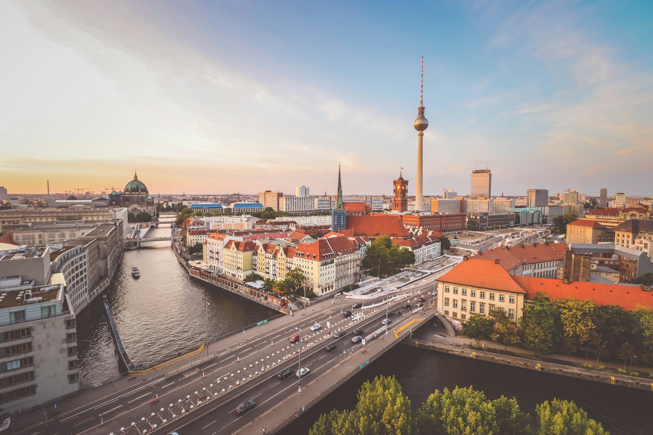 City skyline featuring the Berlin TV Tower, churches, and river with surrounding buildings at sunset.