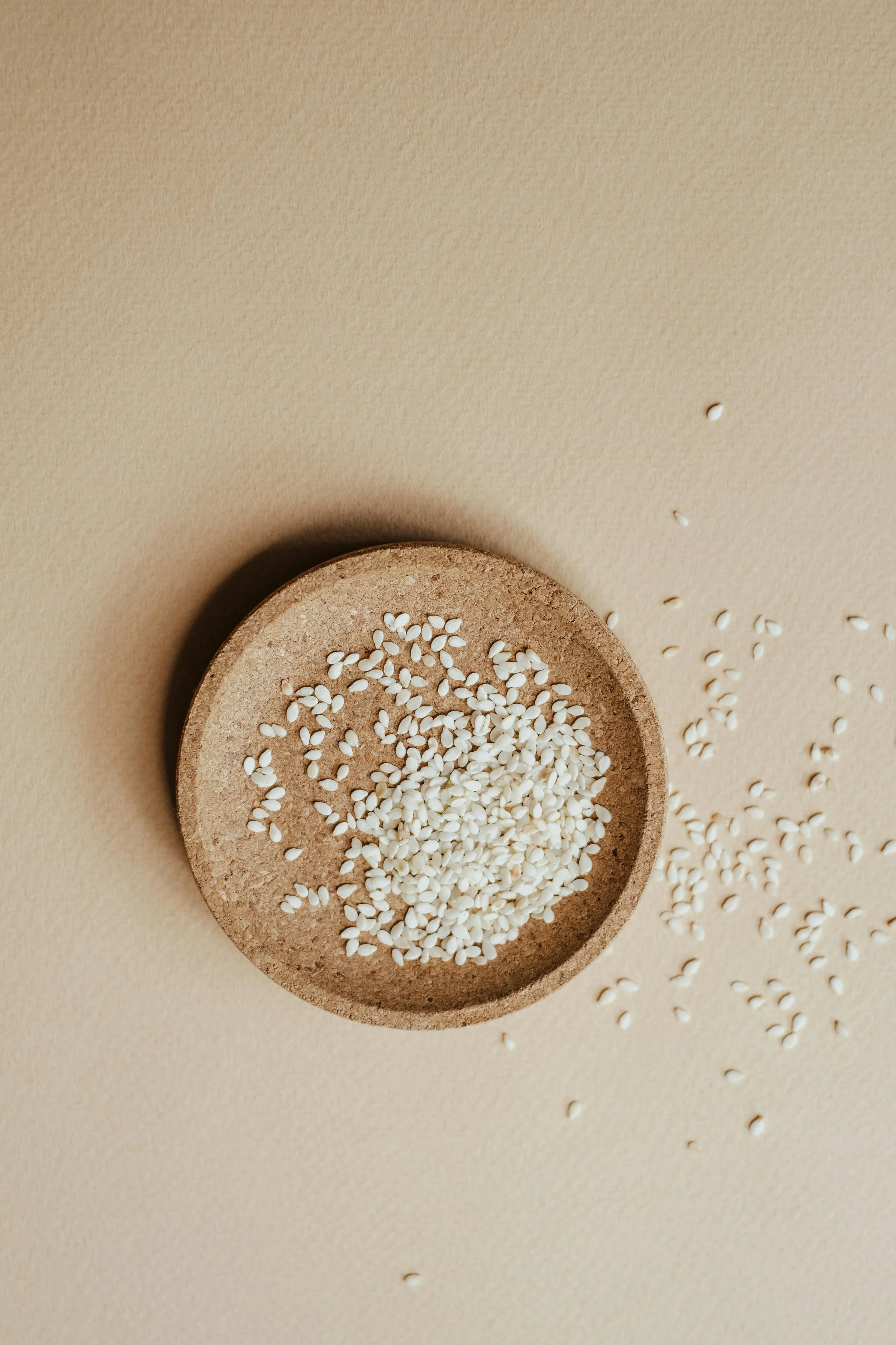 White sesame seeds in a small, round, light brown dish on a beige surface, with some seeds spilled outside the dish.