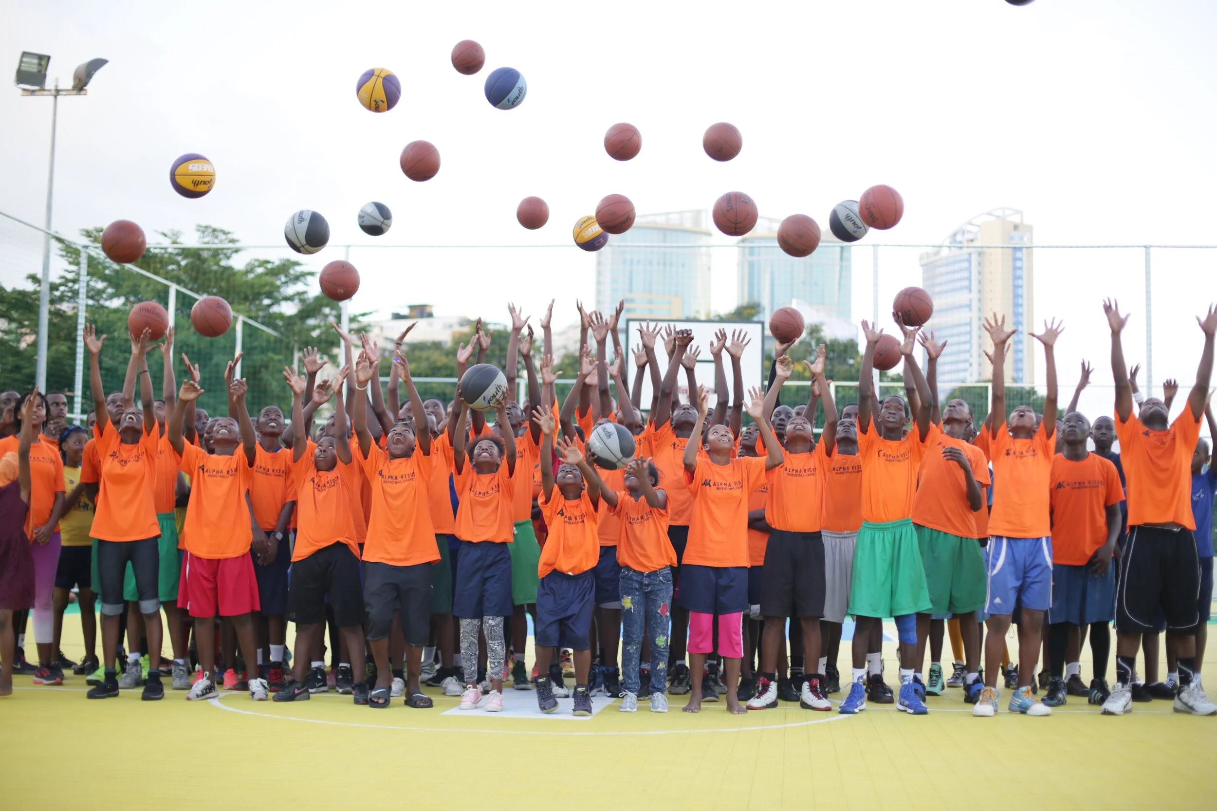 Group of children wearing orange shirts on a basketball court, tossing basketballs and volleyballs into the air.