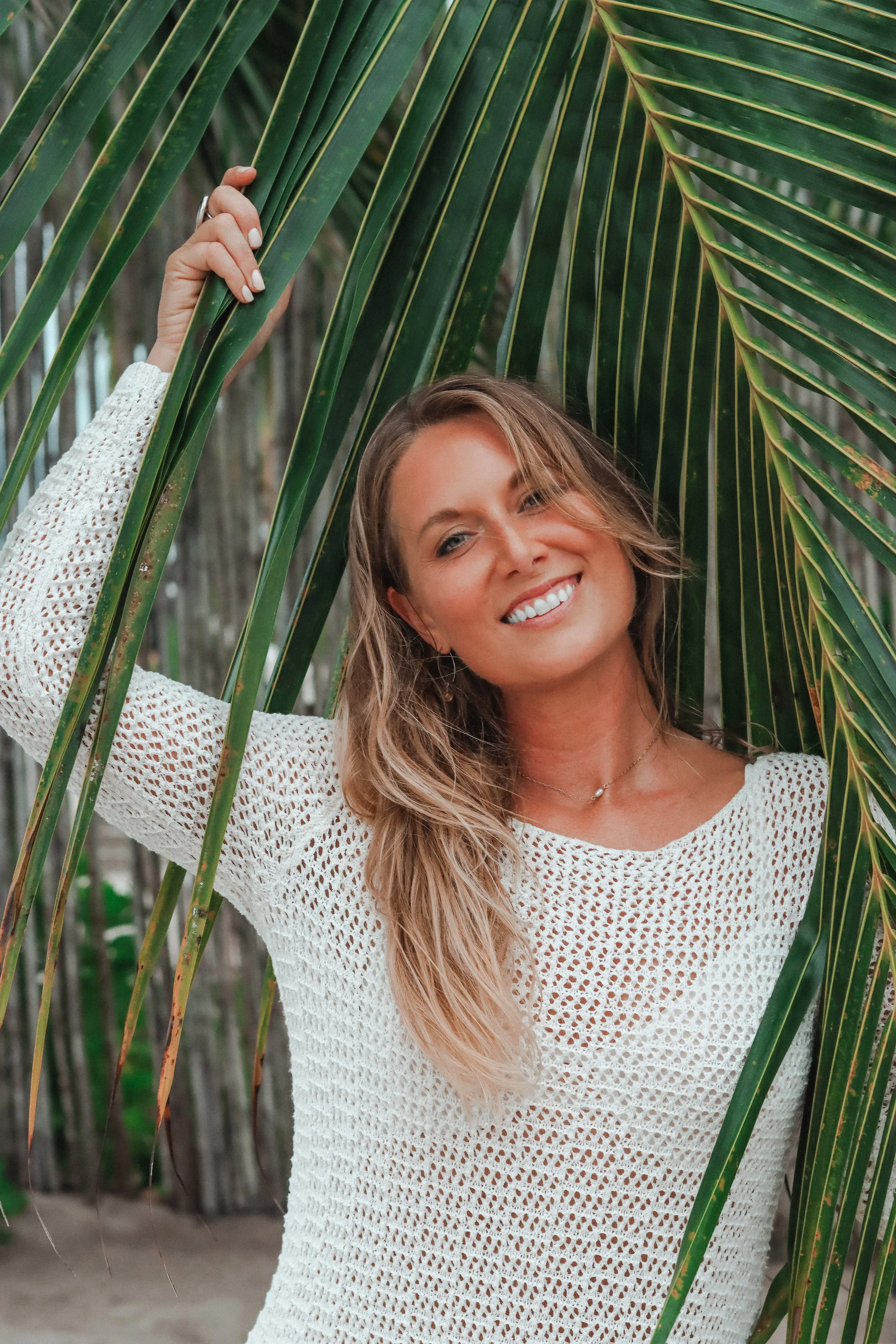 A woman smiling while holding a large green palm leaf in front of a background of palm trees, wearing a white crocheted top.