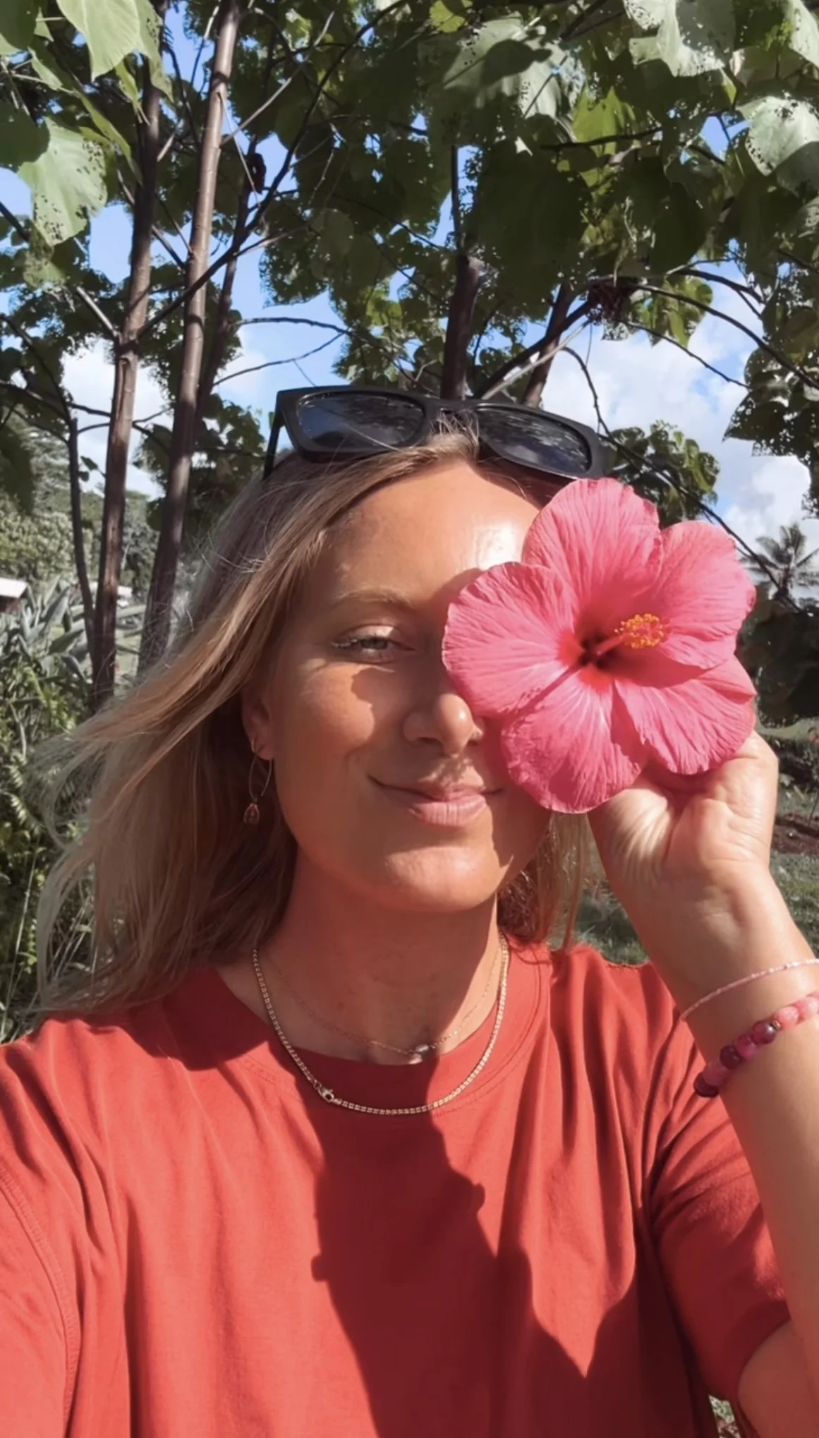 A woman holding a pink hibiscus flower in front of her face outdoors with trees and blue sky in the background.
