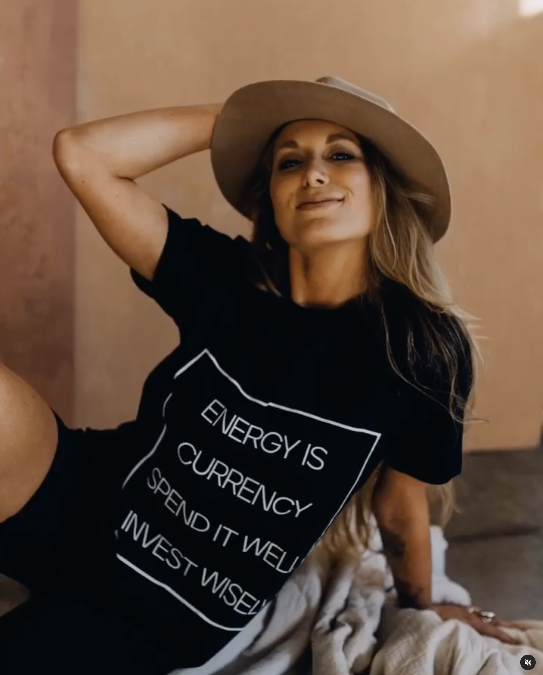 A woman with long, wavy hair and tan skin wearing a beige wide-brimmed hat and a black T-shirt with a message, sitting on a beige blanket, smiling and looking confident.