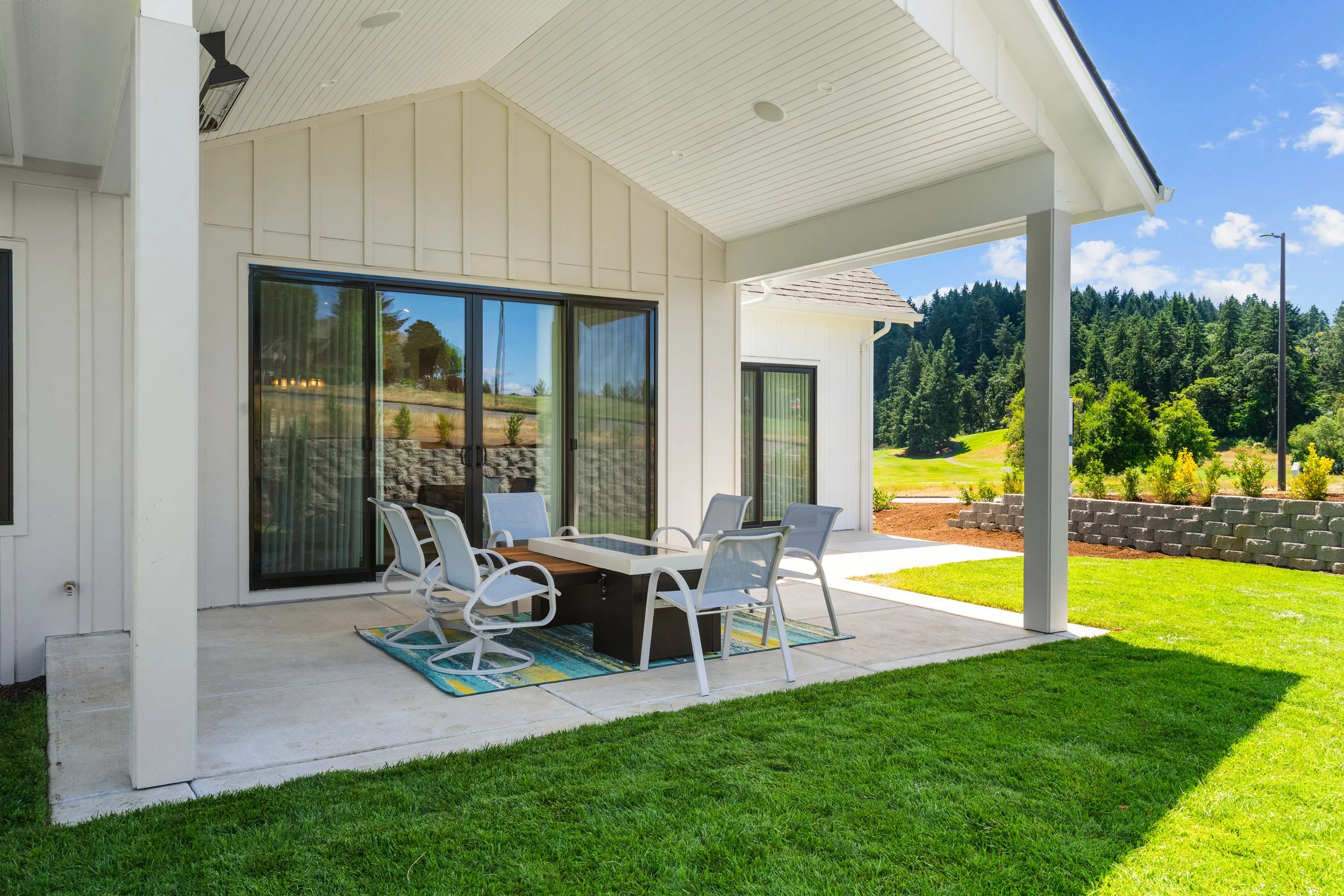 A house patio with glass sliding doors, an outdoor dining table surrounded by white chairs, a colorful outdoor rug, and lush green lawn with trees in the background under a sunny blue sky.