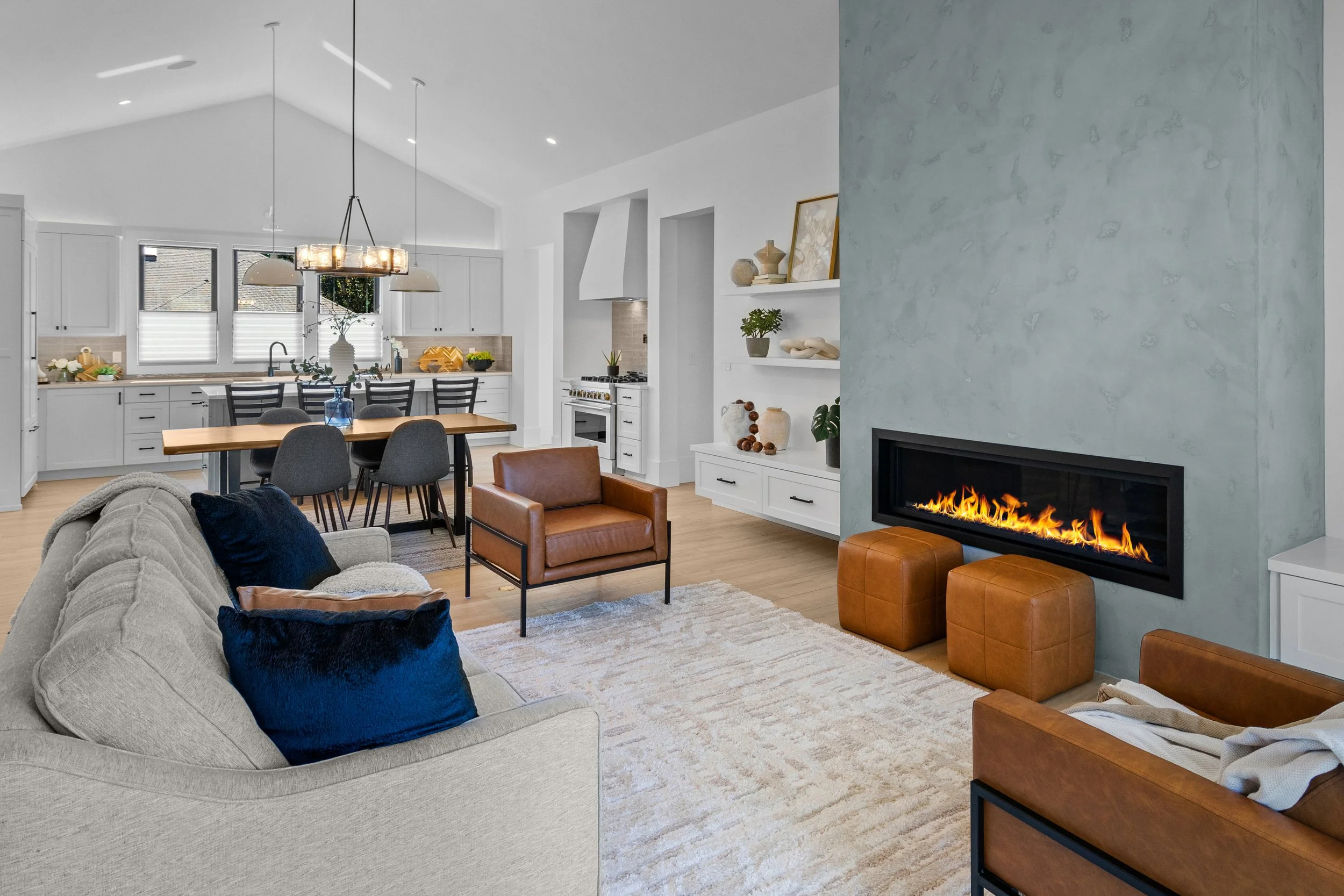 Open-concept living room and kitchen with white walls, light hardwood floors, modern furniture and a fireplace with orange flames. The kitchen has white cabinets, a wooden dining table with dark chairs, and a chandelier above.