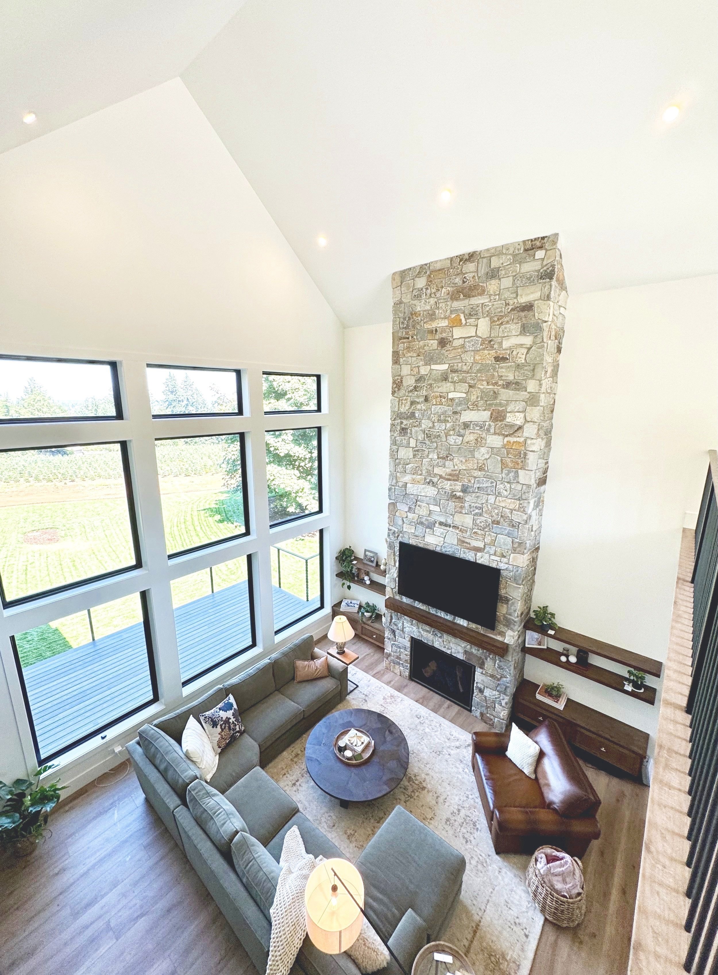 This image shows a spacious living room with a high vaulted ceiling, large windows, a stone fireplace, a gray sofa, a brown leather armchair, a round coffee table, and built-in wall shelves with decorative items and plants.