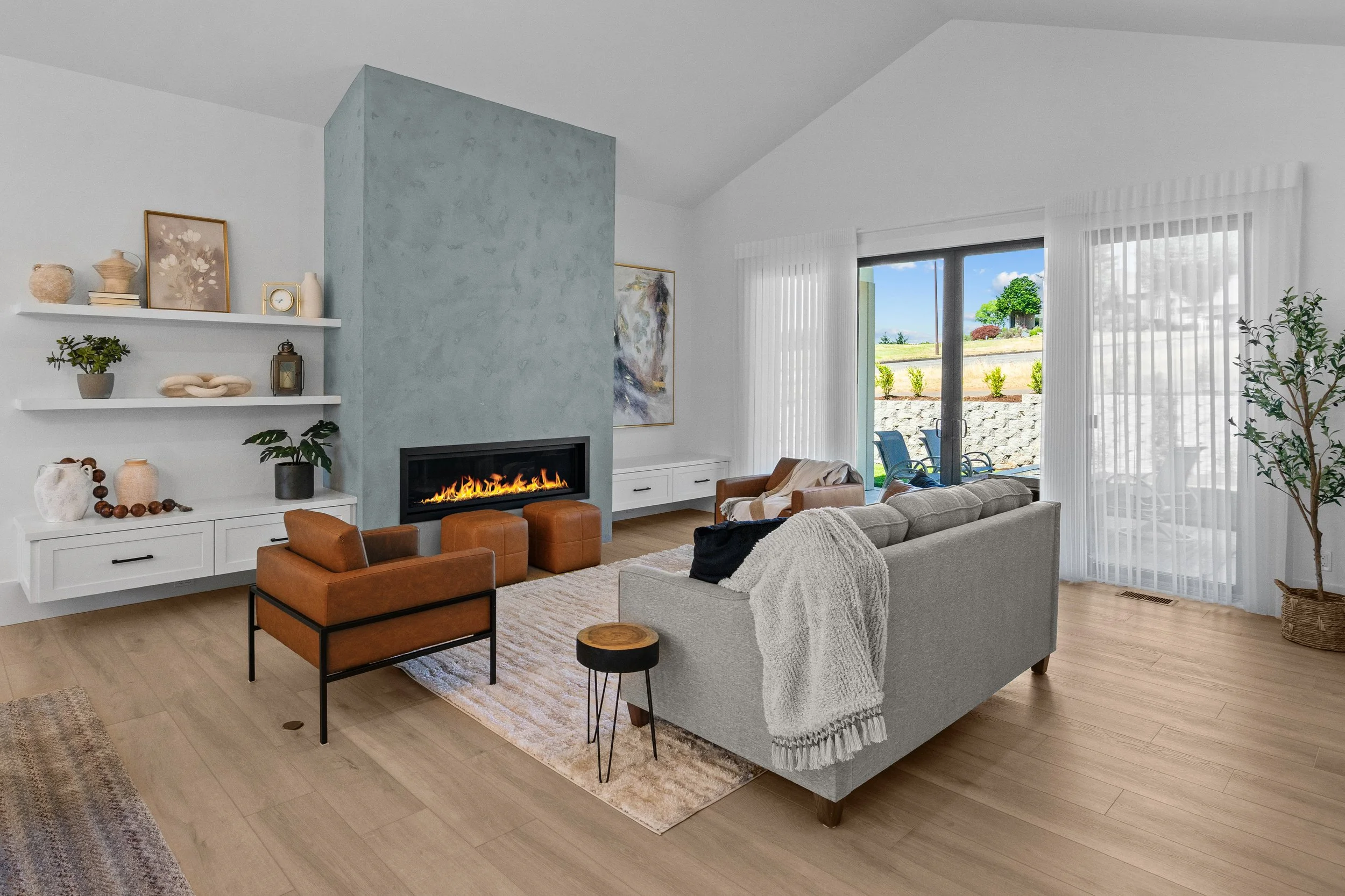 Living room with a modern electric fireplace, white walls, and large glass doors leading outside. The room has a gray sofa with a blanket, a brown armchair, and decorative items on white shelves, including plants and art pieces. Outdoor patio furnitu