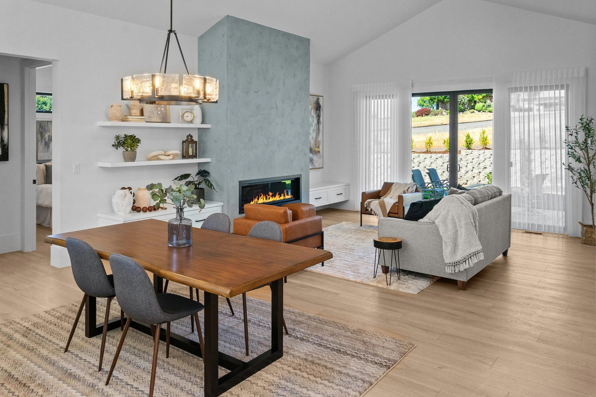 A modern living room with a dining table, gray chairs, a cozy gray sofa, a fireplace, open windows with a view of a garden, white shelves with decorative items, and a large chandelier.