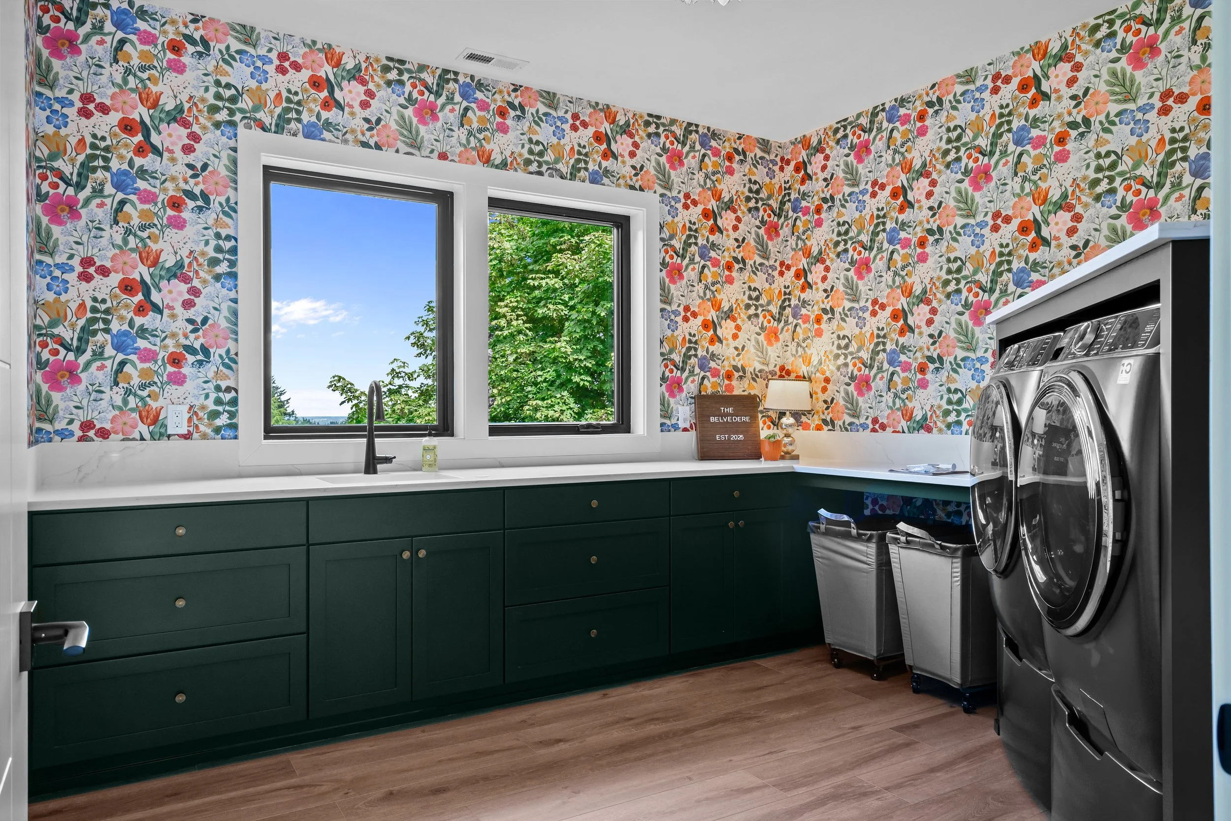 A laundry room with floral wallpaper, dark green cabinets, a window showing green trees and blue sky, a white countertop, and two stacked front-loading washing machines.