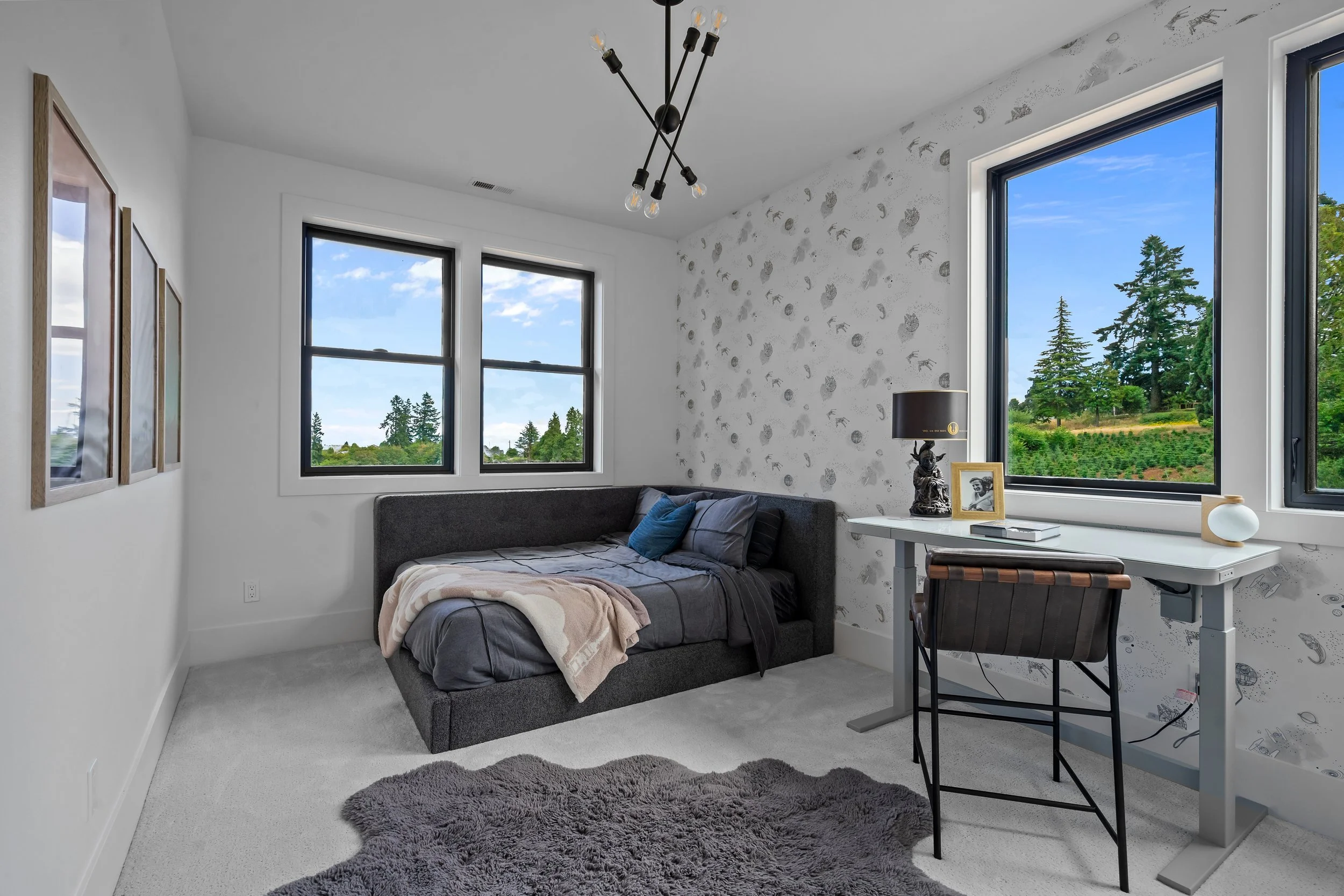 Interior of a bedroom with white walls, two large windows showing green trees outside, a gray bed with blankets and pillows, a shaggy gray rug, a desk with a lamp and framed photograph, and a modern black chandelier hanging from the ceiling.