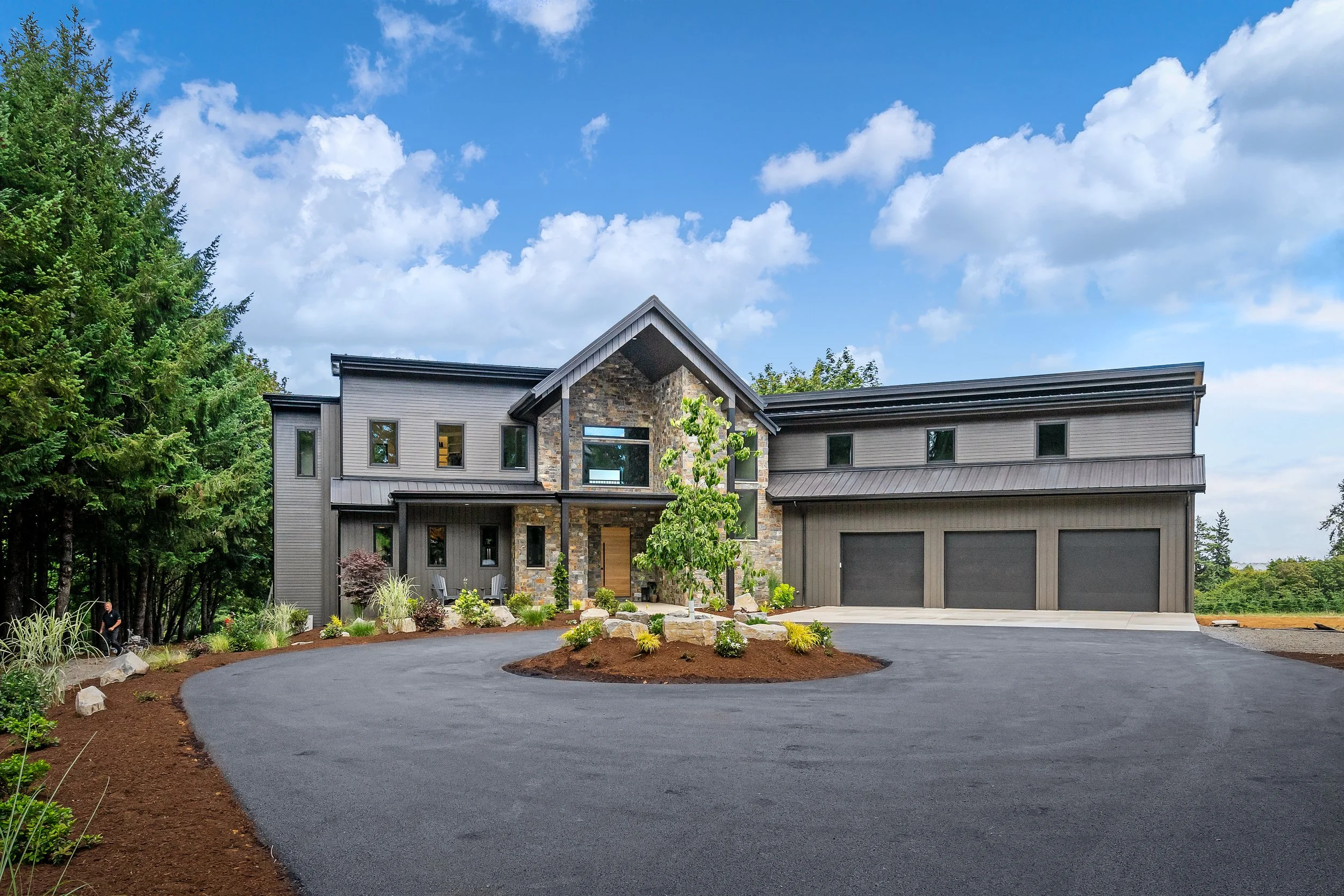 A modern house with gray siding and stone accents, surrounded by landscaped yard and trees, under a bright blue sky with puffy clouds.