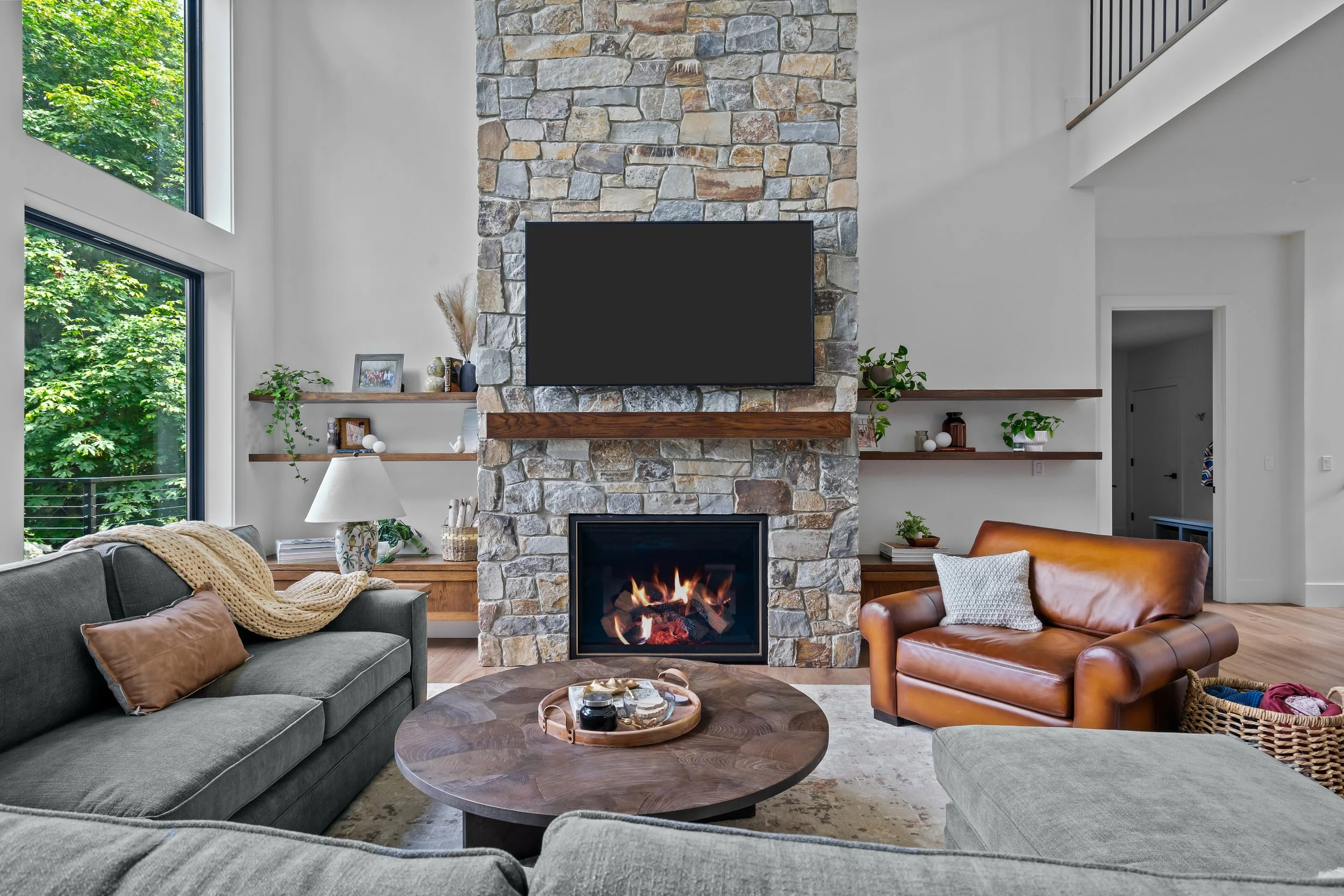 Living room with two sofas, one gray and one tan leather, a round wooden coffee table with a tray, a fireplace, and a stone chimney with a mounted flat-screen TV. Large windows with greenery outside.