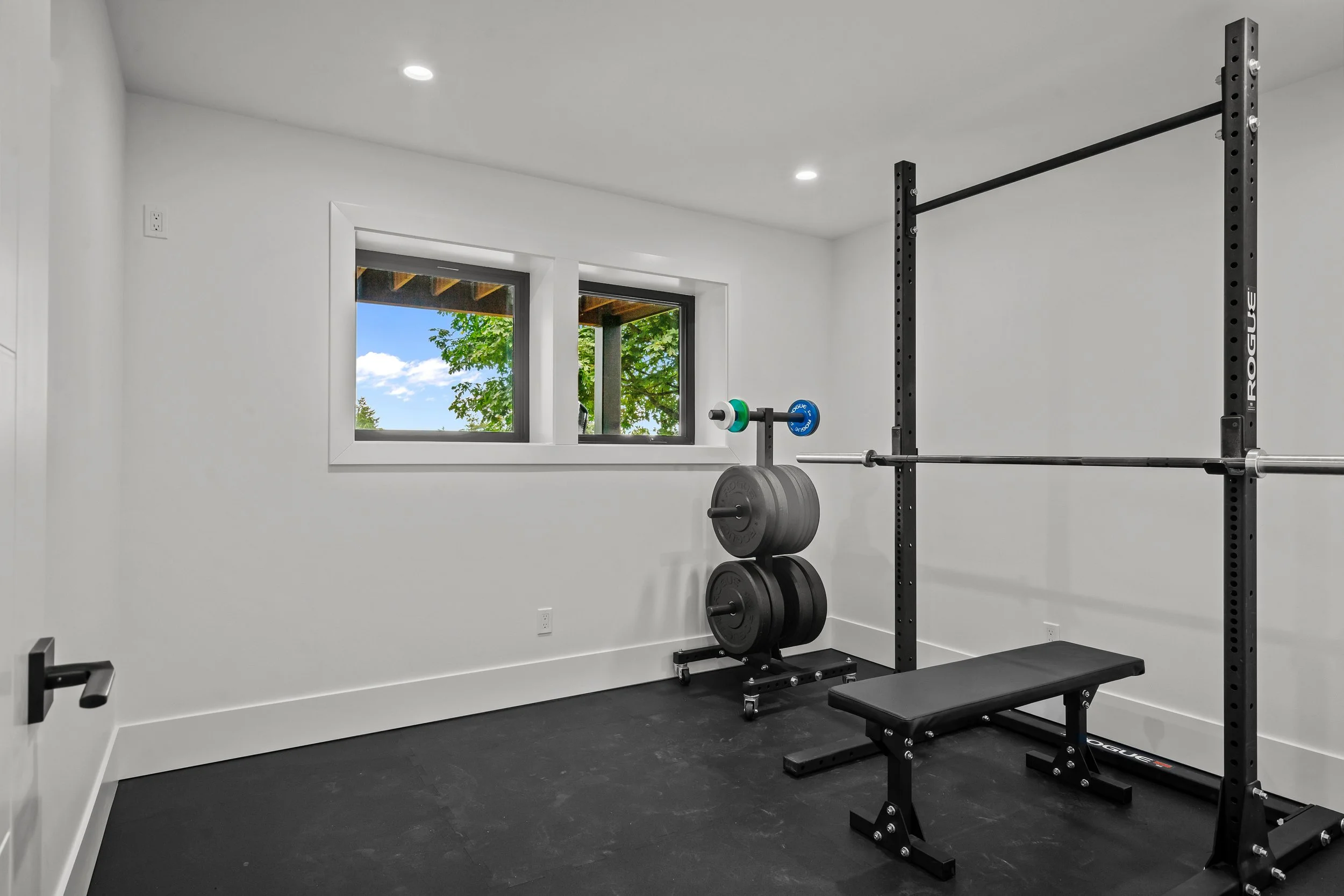 Home gym with black power rack, barbell with weights, and a weight bench in a room with white walls, two windows, and black flooring.