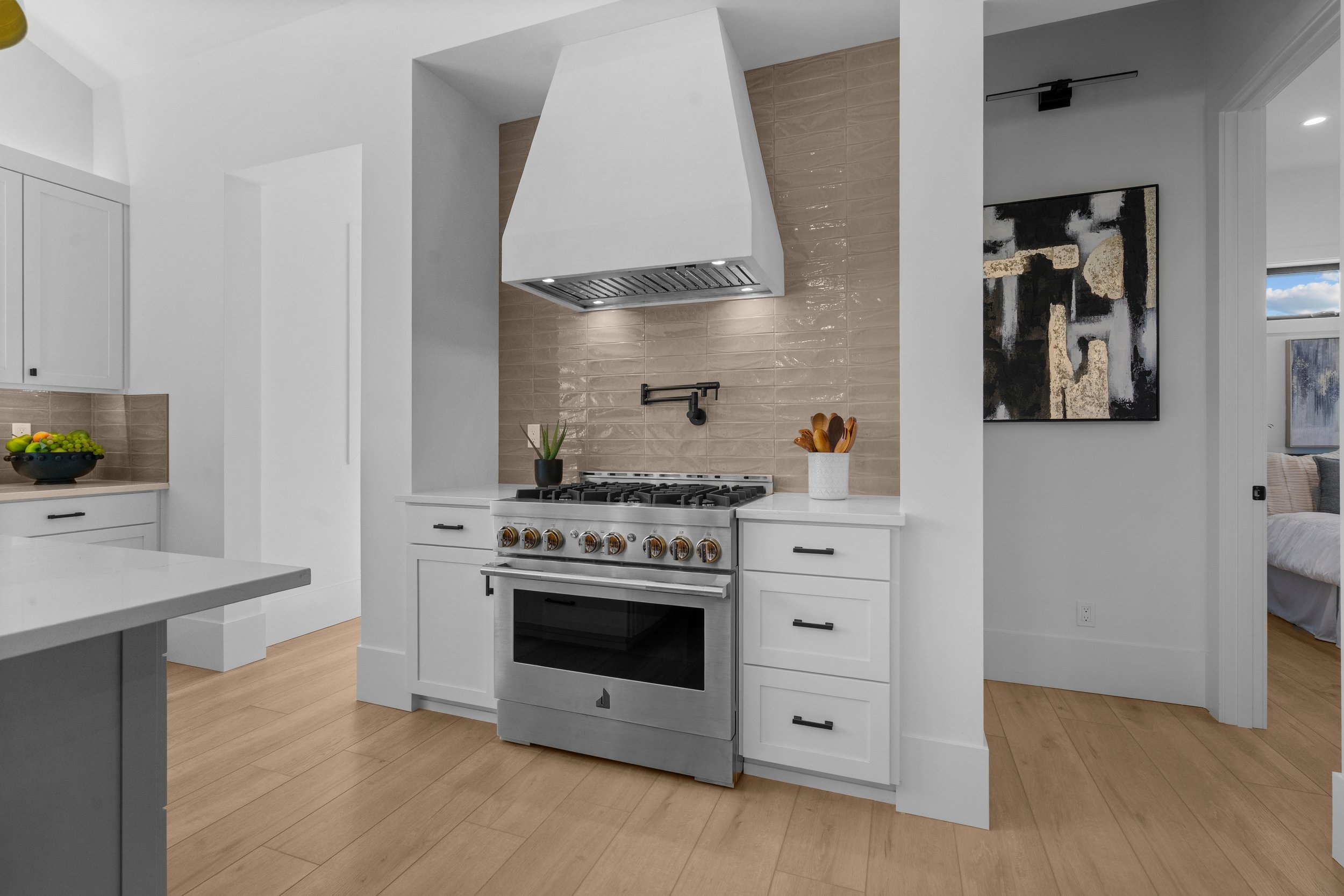 Modern kitchen with white cabinets, beige tiled backsplash, stainless steel stove with gas burners, white range hood, and light wooden flooring.