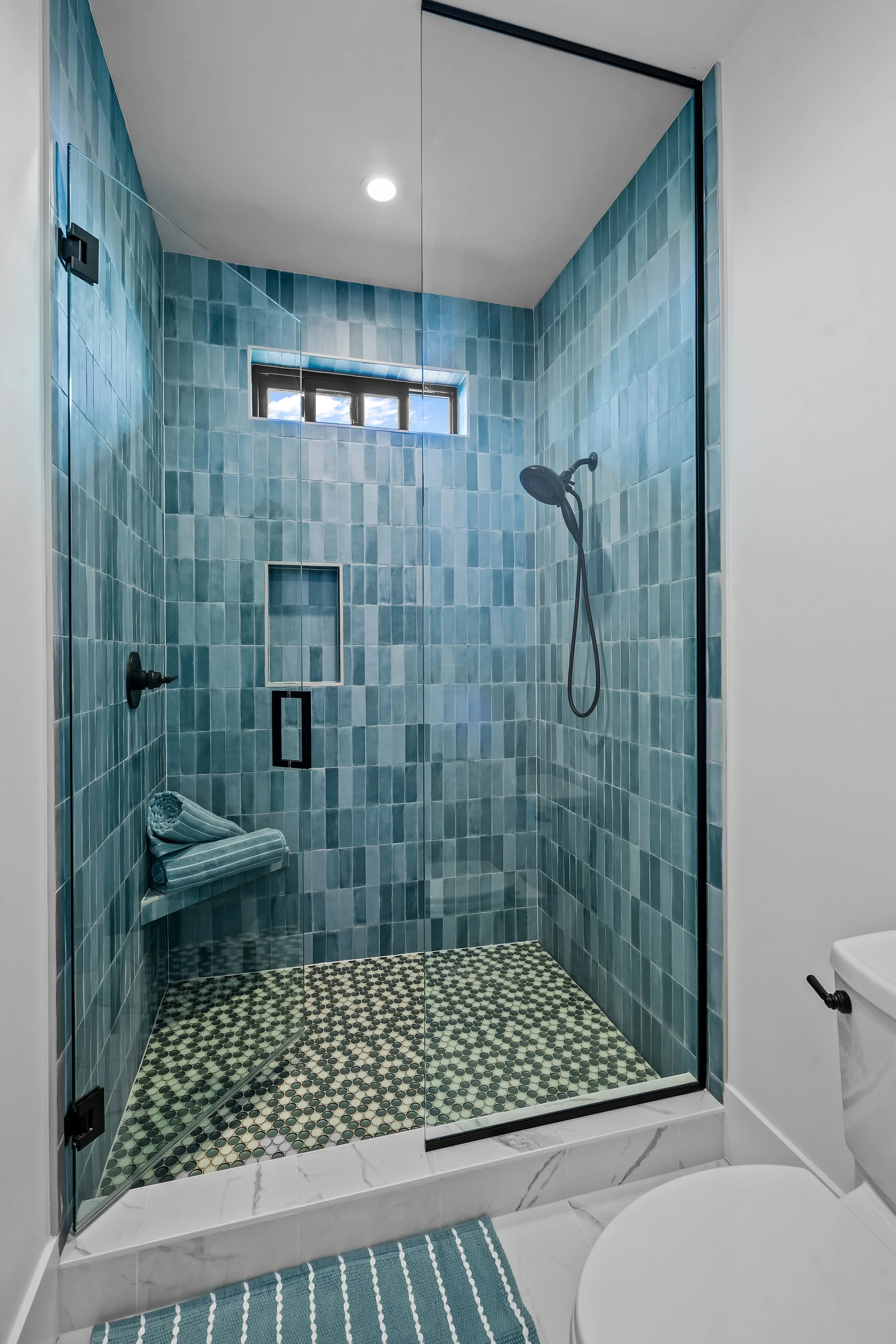 Modern walk-in shower with blue tiles, a small window showing a partly cloudy sky, black fixtures, and a glass door with black hinges, located in a bathroom with a white marble floor and a striped blue rug.
