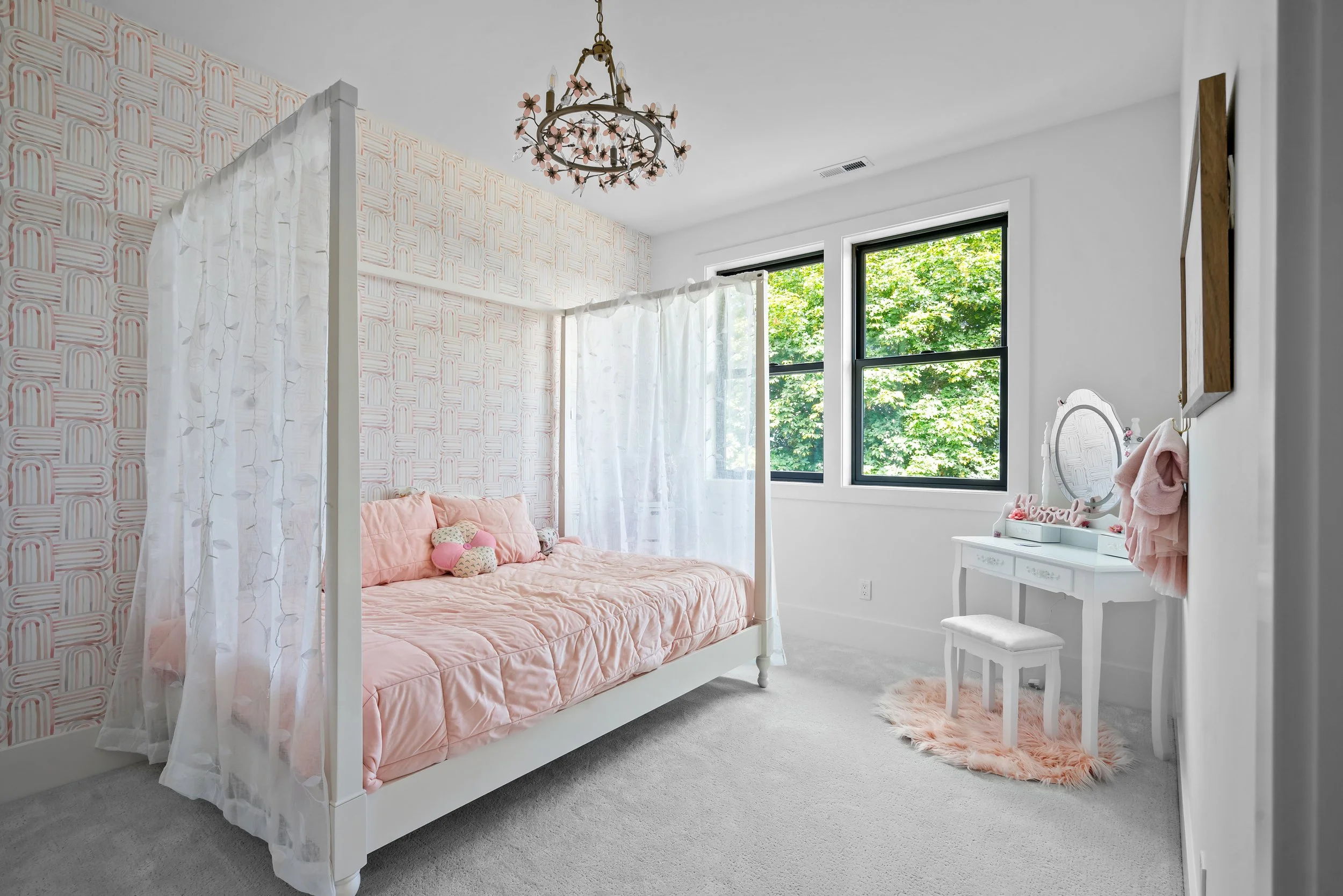 A cozy children's bedroom with a white canopy bed dressed in pink bedding, a pink plush toy, and pastel decor. The room features a vanity table, a fluffy pink rug, a chandelier with floral accents, and large windows with a view of green trees outside