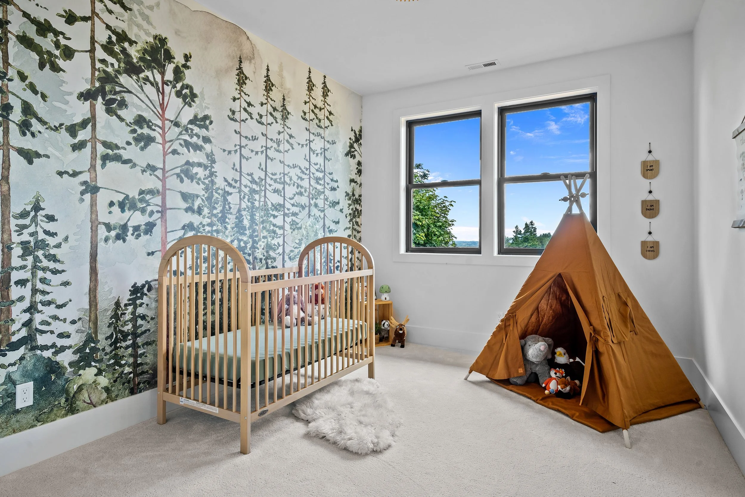 A nursery with a forest-themed wallpaper, a wooden crib with stuffed animals, a small wooden nightstand with a toy, a cozy tent with stuffed animals inside, and two windows showing a clear blue sky and green trees outside.