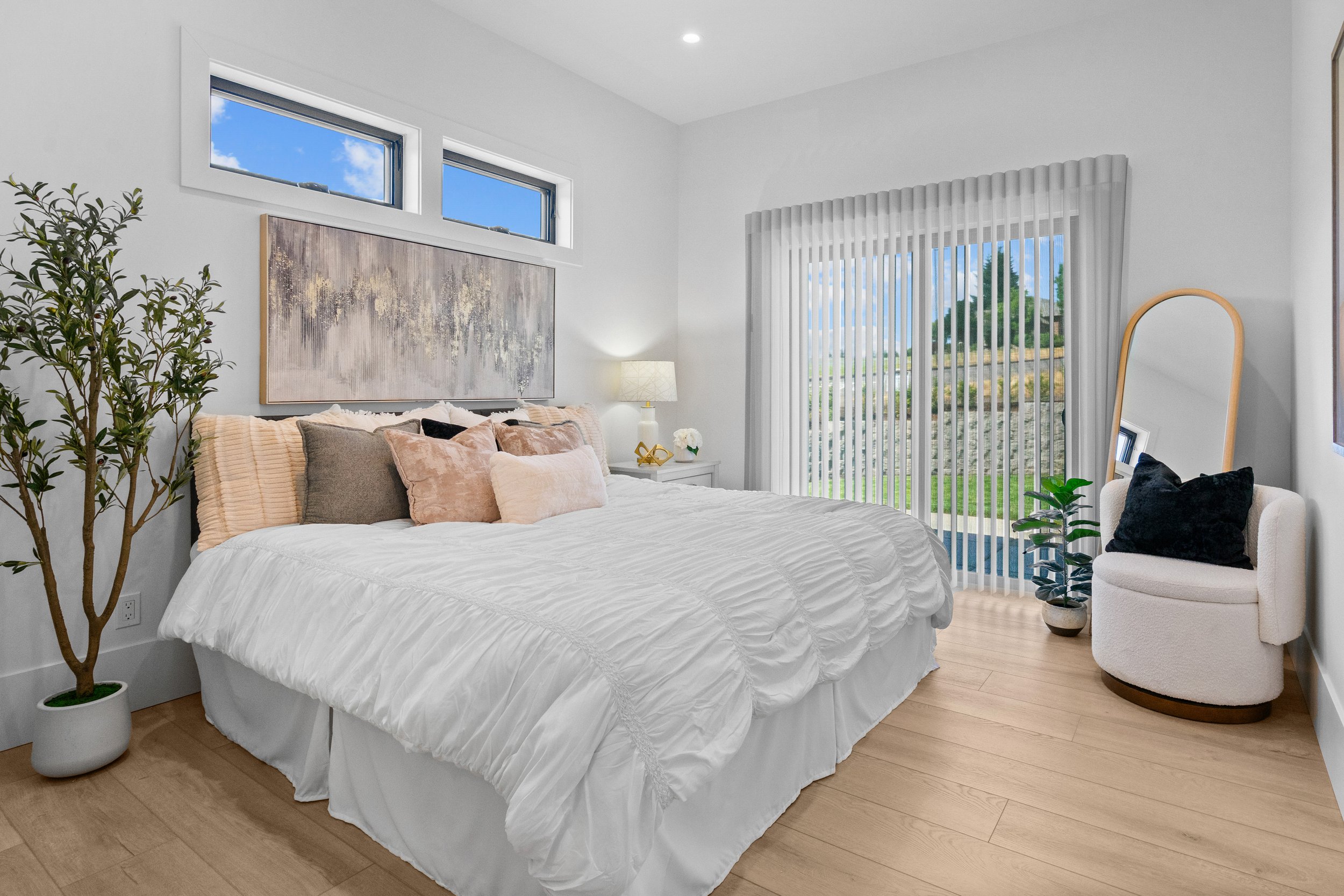 A neatly made bed with white bedding and multiple throw pillows in shades of beige, pink, and black in a modern bedroom with large windows, a white bedside table with a lamp and and vertical blinds covering a sliding glass door.