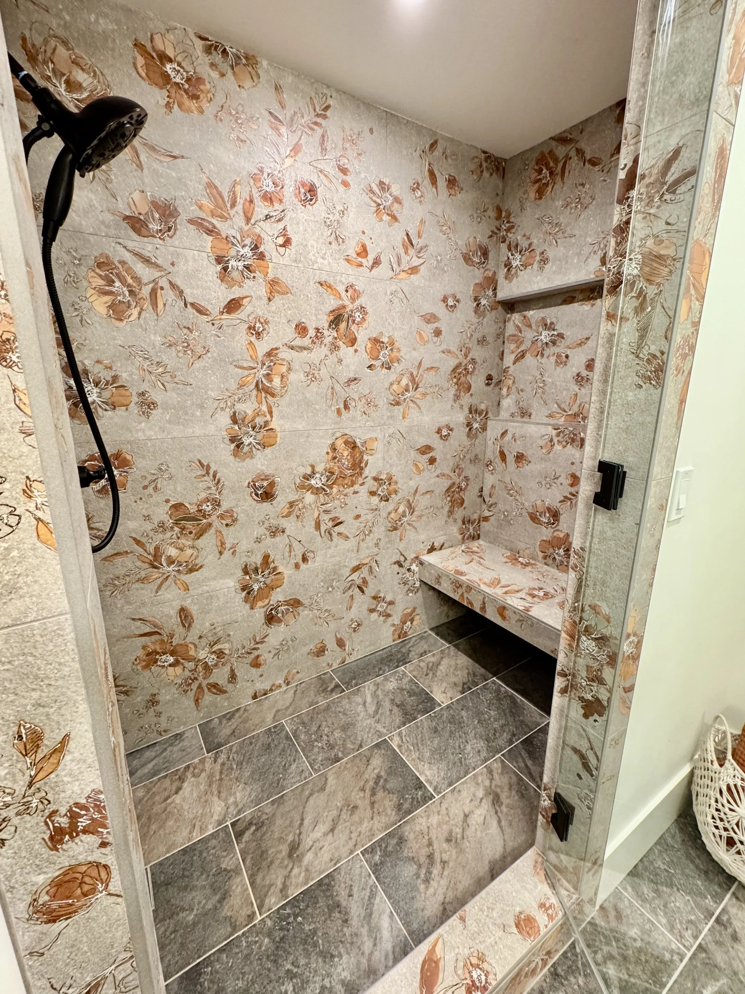 Bathroom shower area with floral patterned tiles on the walls, a dark shower head, and a small marble ledge. The floor has grey tiles and a mirror on the right side reflecting part of the space.