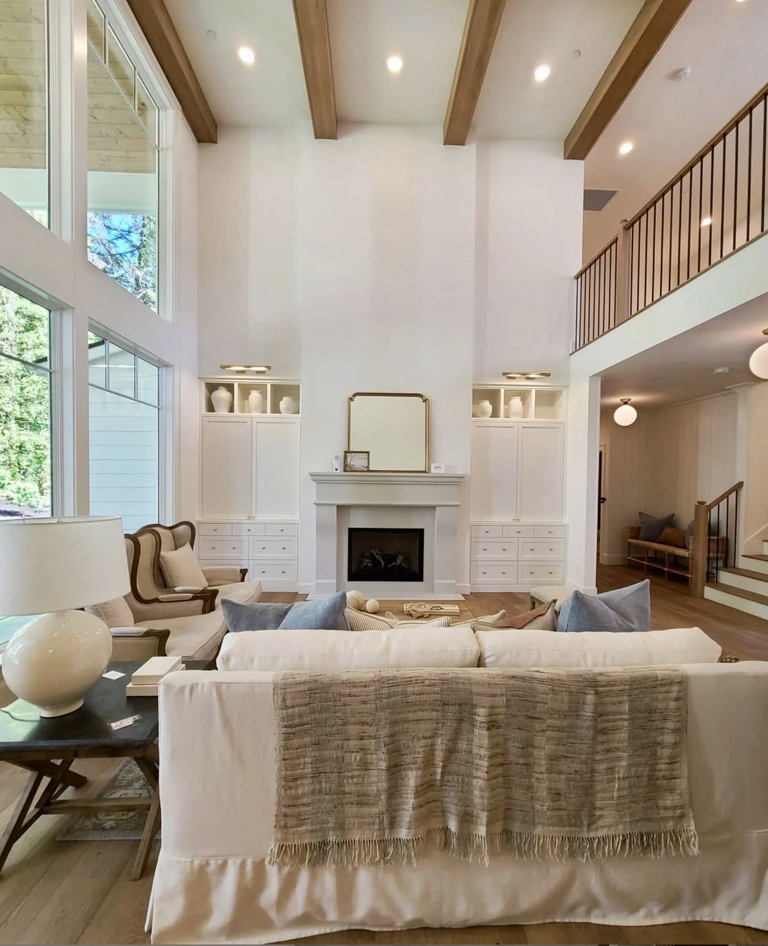 Living room with high ceilings featuring wood beams. Large windows, white built-in shelves and french stone fireplace.