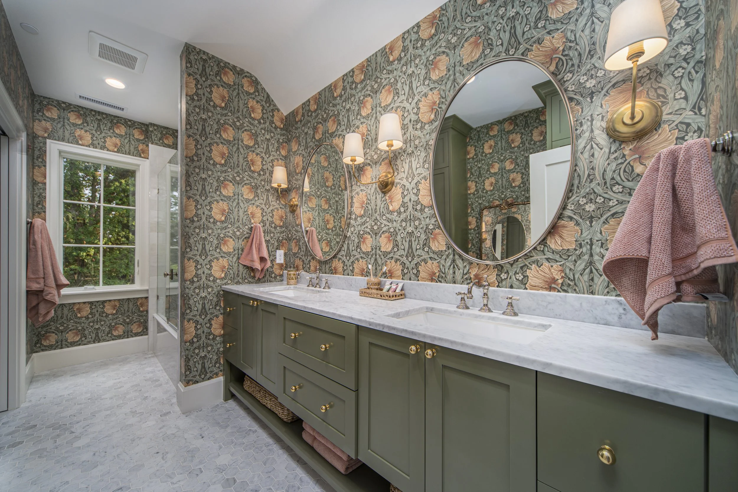 Bathroom with green cabinetry, marble countertop, large oval mirror and floral wallpaper.