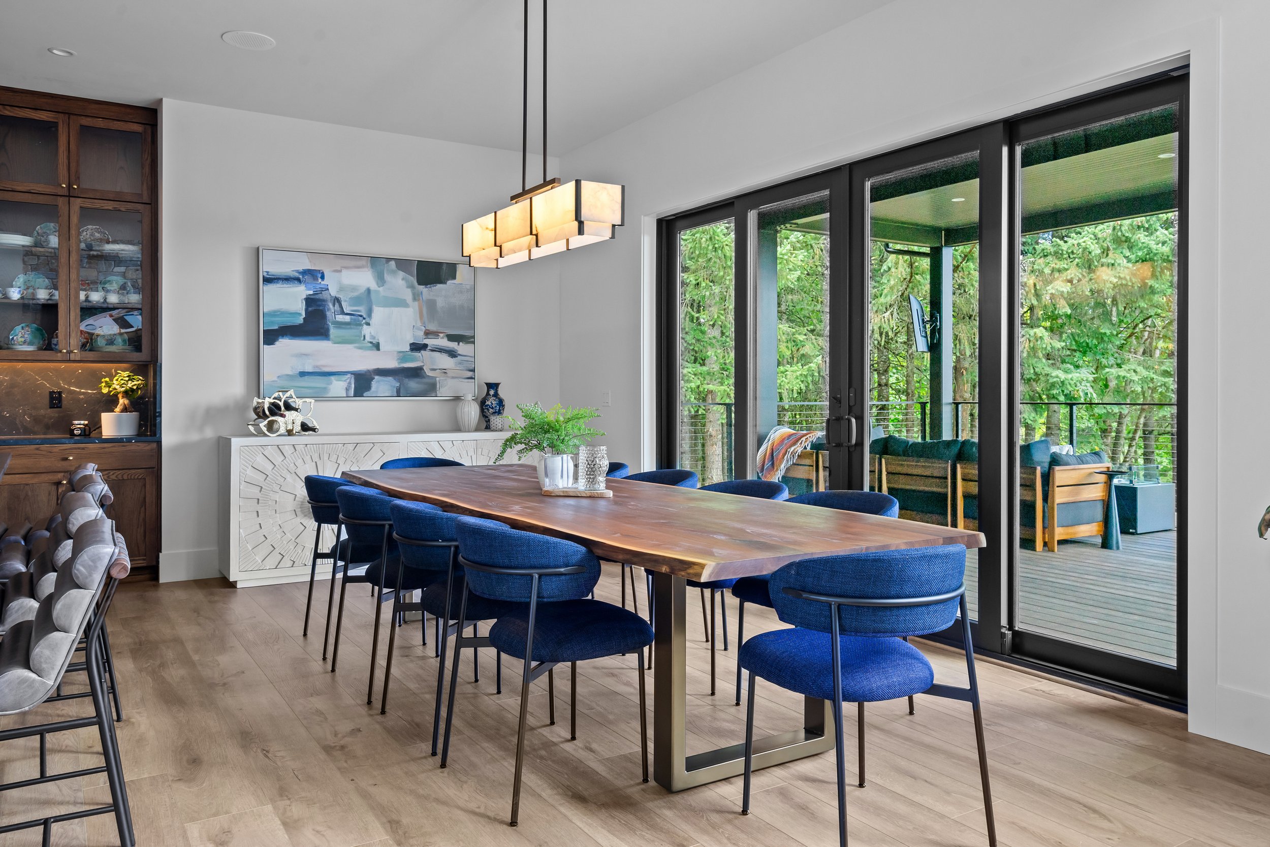A modern dining room with a wooden table surrounded by blue upholstered chairs, large sliding glass doors leading to a balcony with green trees outside, artwork on the wall, a chandelier, and a built-in wooden cabinet with decorative items.