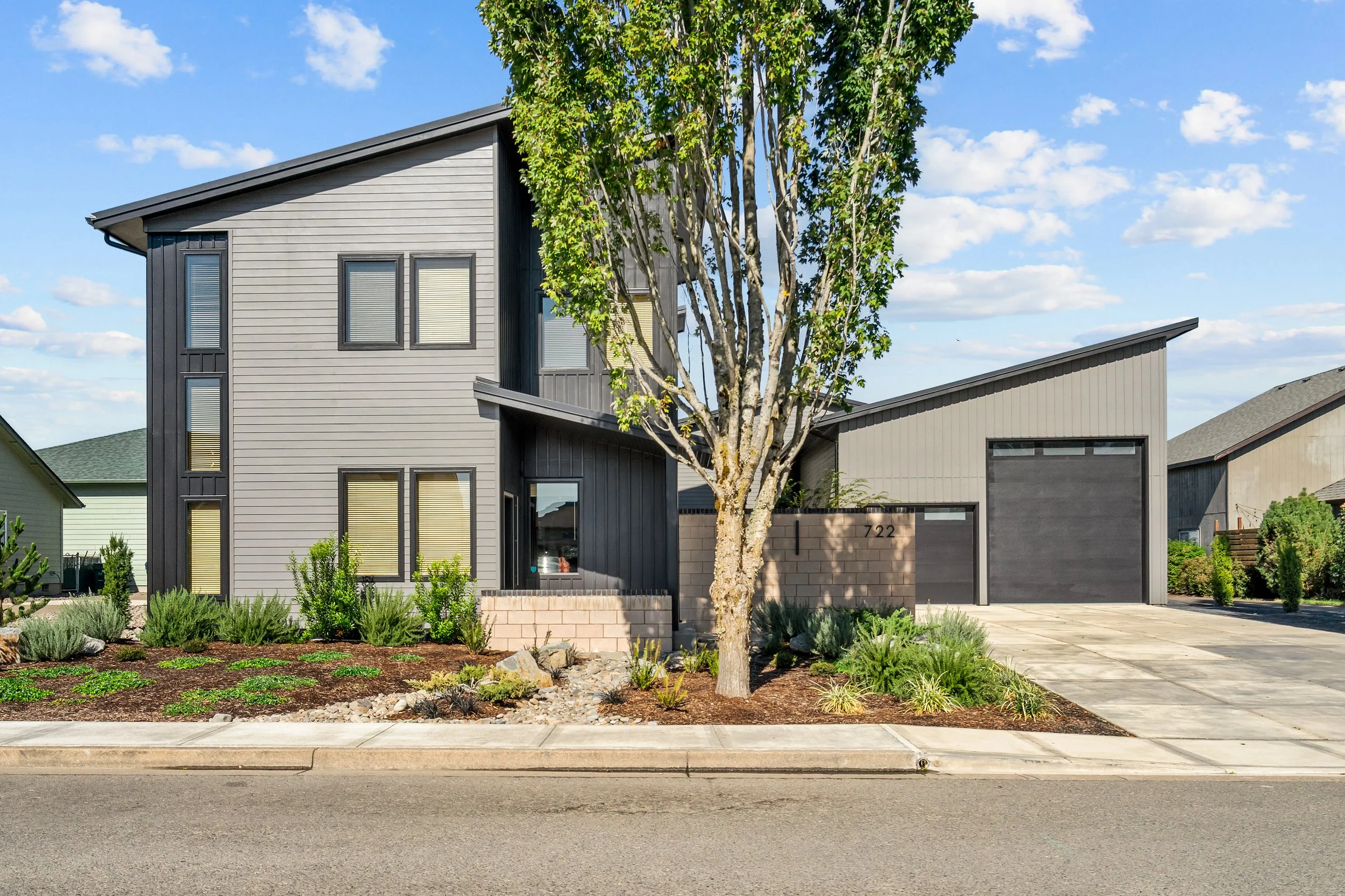 Modern gray house with black window frames, a slanted roof, a small front yard with bushes, a tree, and a concrete driveway, under a blue sky with scattered clouds.