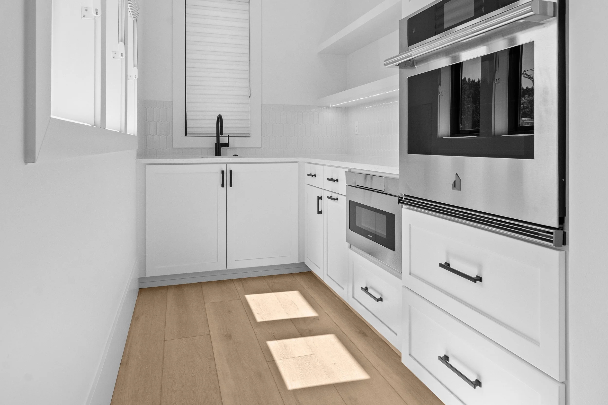 A butlers pantry with white cabinets, black handles, a black sink faucet, a window, and stainless steel appliances, including an oven and microwave, with natural light coming through the window.