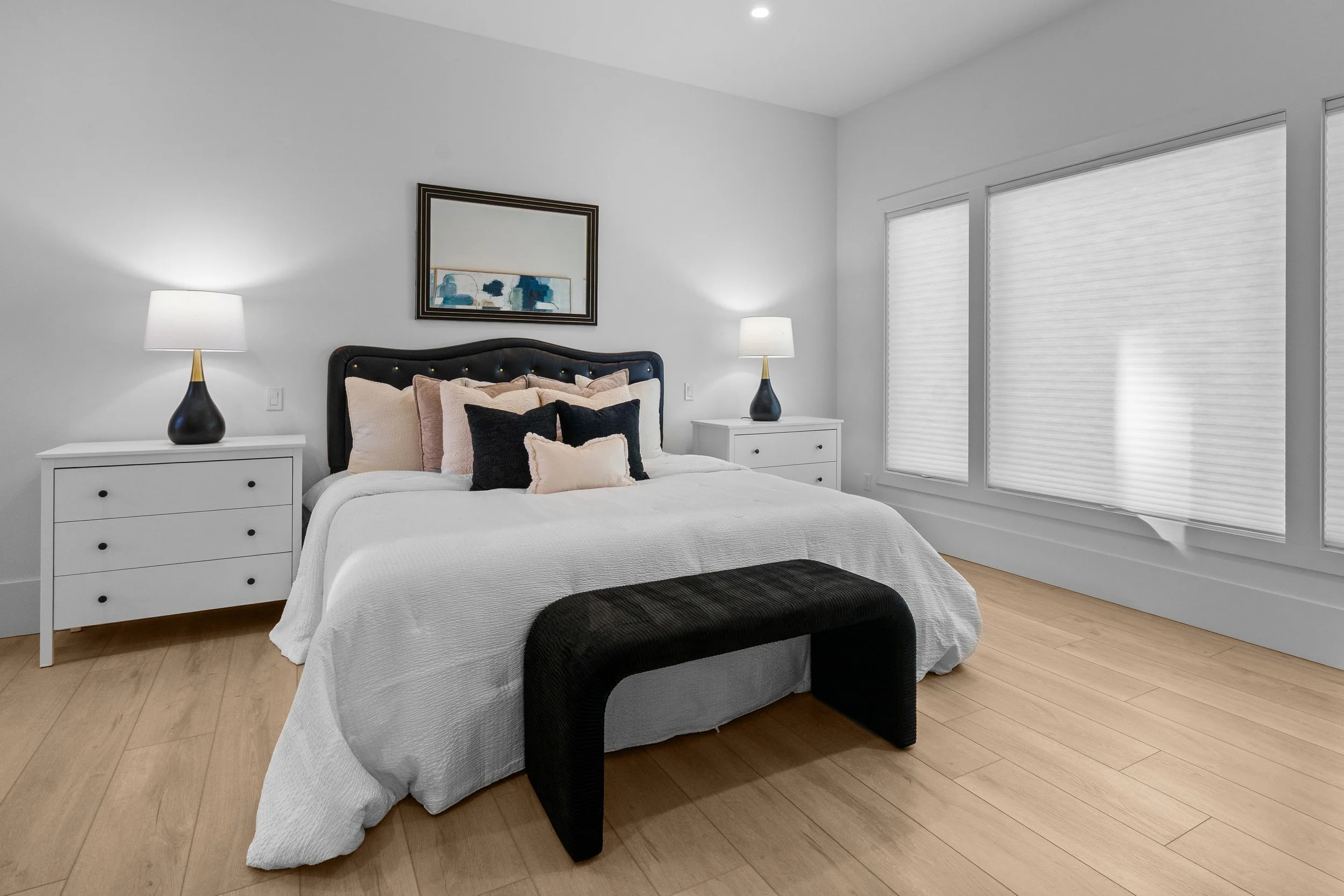 A modern bedroom featuring a bed with a white bedspread, multiple pillows, and a black headboard. Flanking the bed are white nightstands with black and gold lamps. The room has light wood flooring and large windows with white blinds, allowing natural