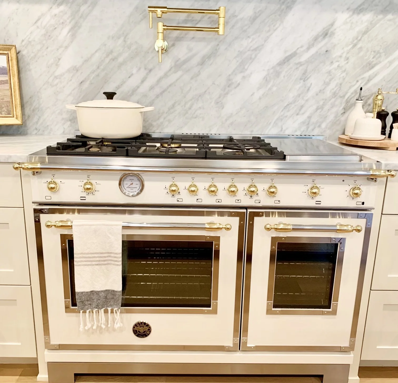 A vintage-style kitchen stove with a white and gold accents, featuring four burners on top and two ovens below. The stove is set against a marble backsplash with a gold pot filler.