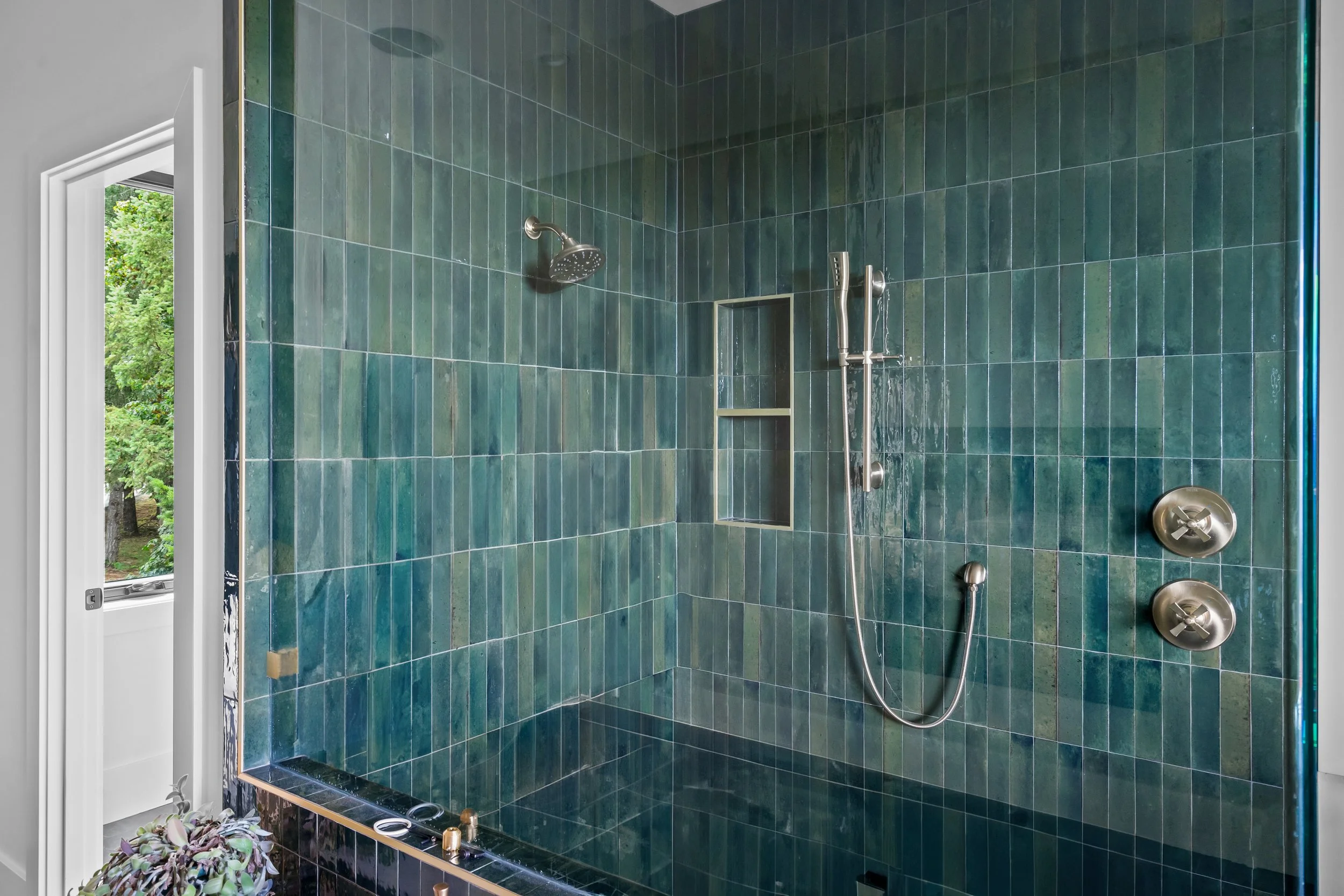 A walk-in shower with dark green tiled walls, a silver rain showerhead, a handheld showerhead on a bar, and a built-in shelf. Part of an open window with views of green trees is visible.