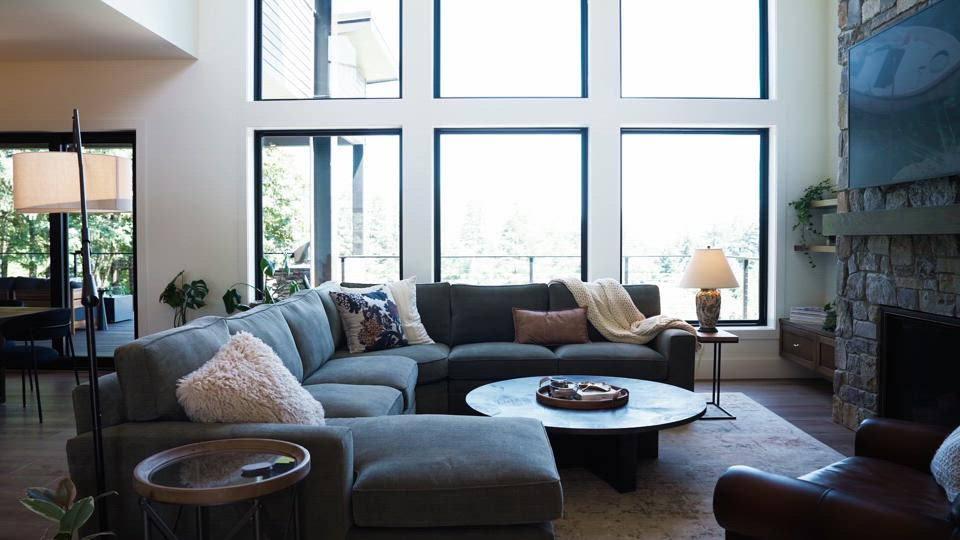 Living room with large windows, a gray sectional sofa, a round coffee table, a lamp on a side table, and decorative pillows.
