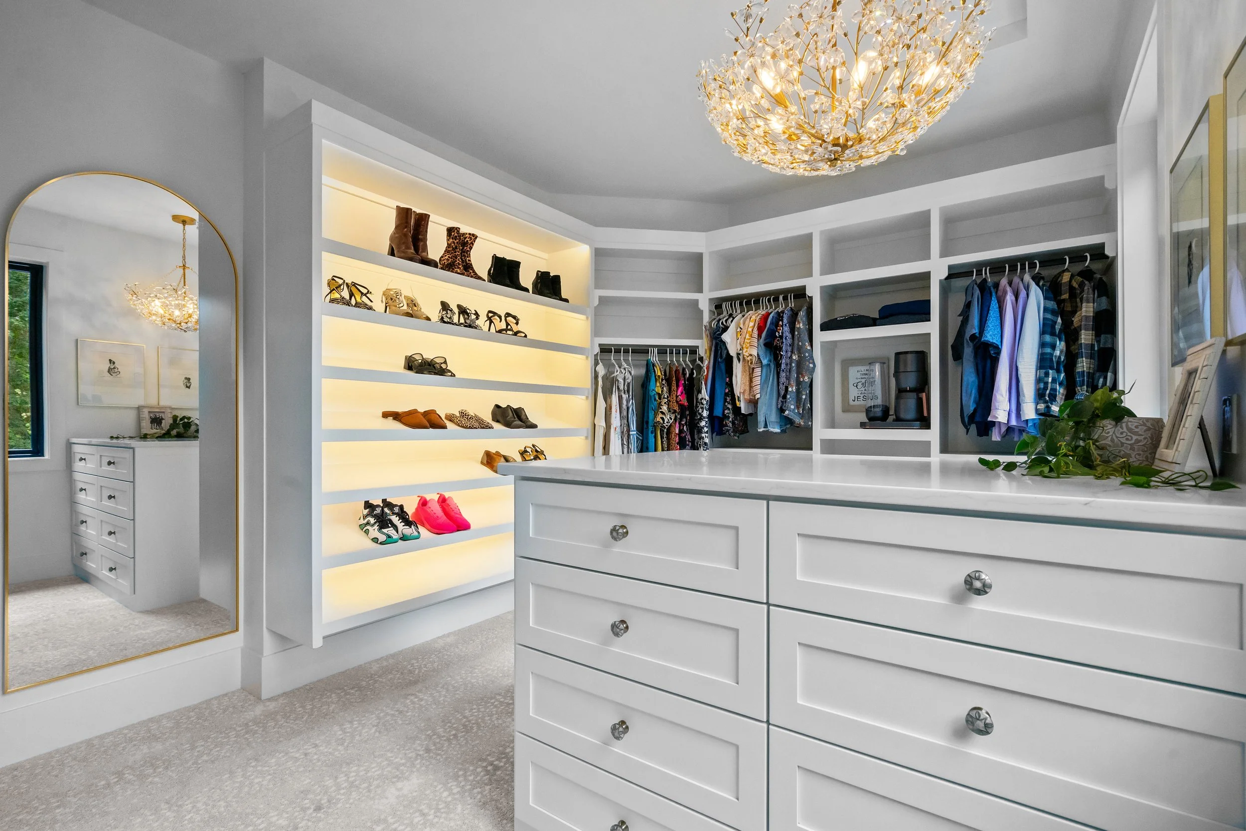 Walk-in closet with white shelving, open clothing racks, a shoe display with illuminated shelves, and a large mirror. Decor includes a chandelier and framed artwork.