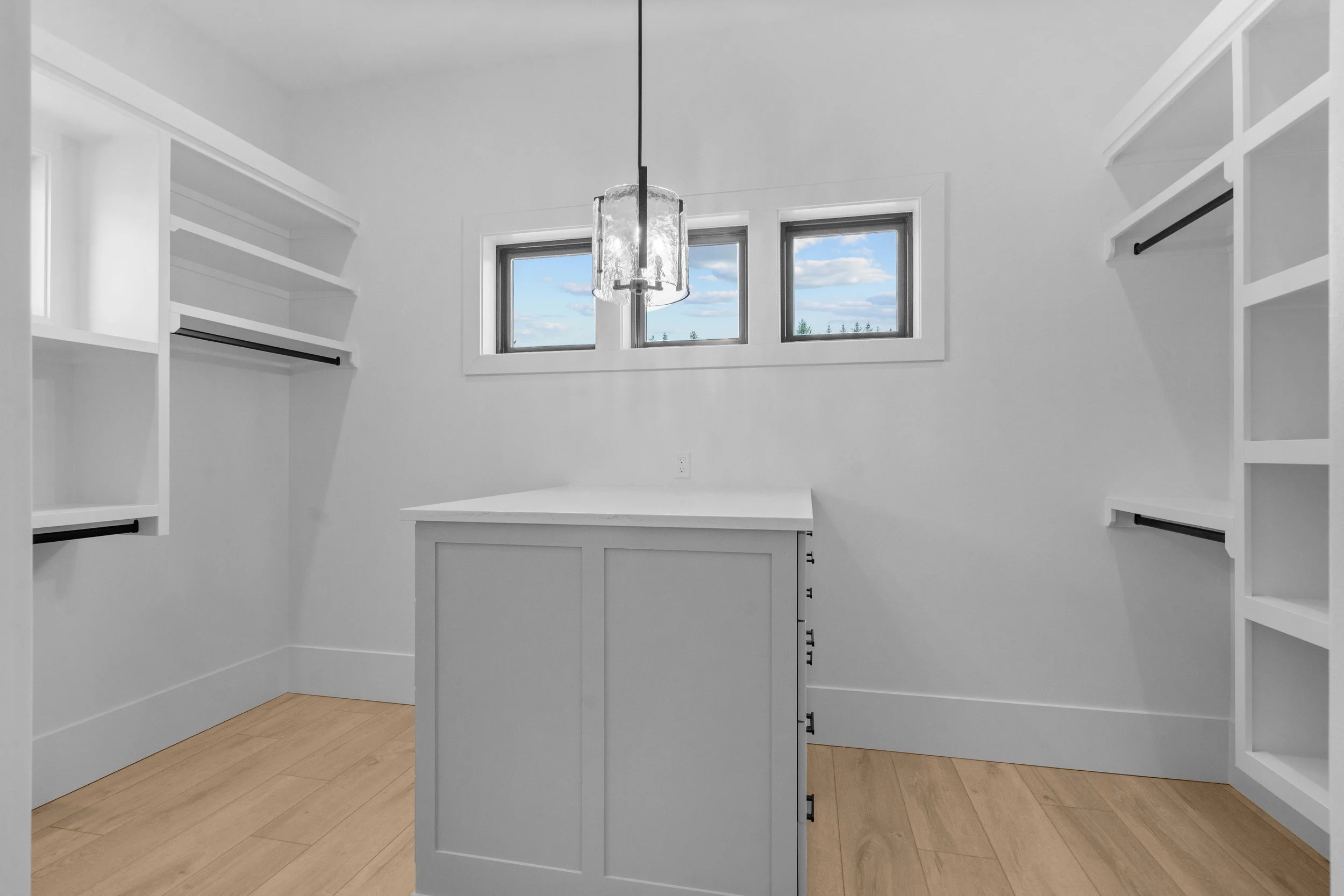 Walk-in closet with white shelves and black rods, a small window showing blue sky and clouds, light wood floor, and a hanging light fixture.