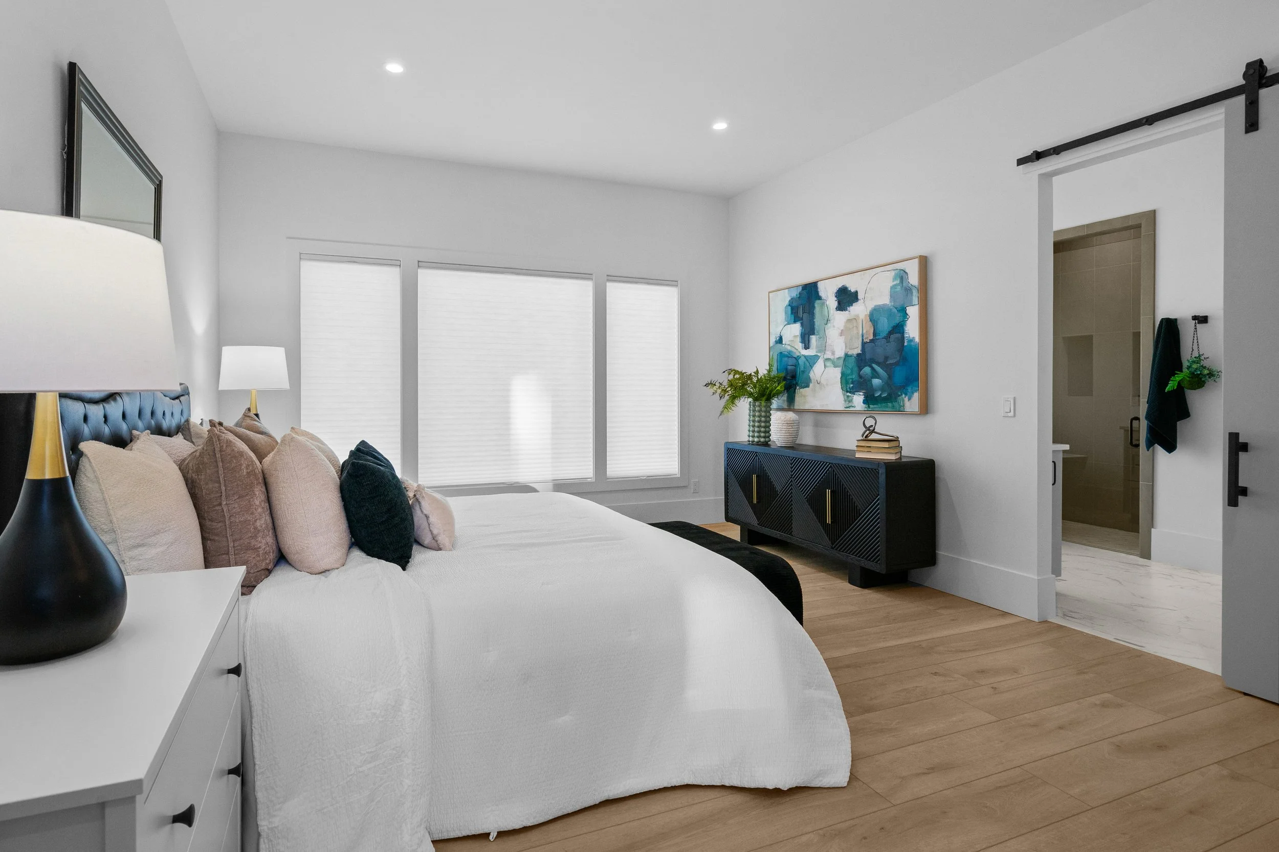 A bedroom with a white bed and a headboard with multiple pillows, a black vase lamp, a blue console table with decor, a large abstract blue and white painting, and a sliding barn door leading to a bathroom.