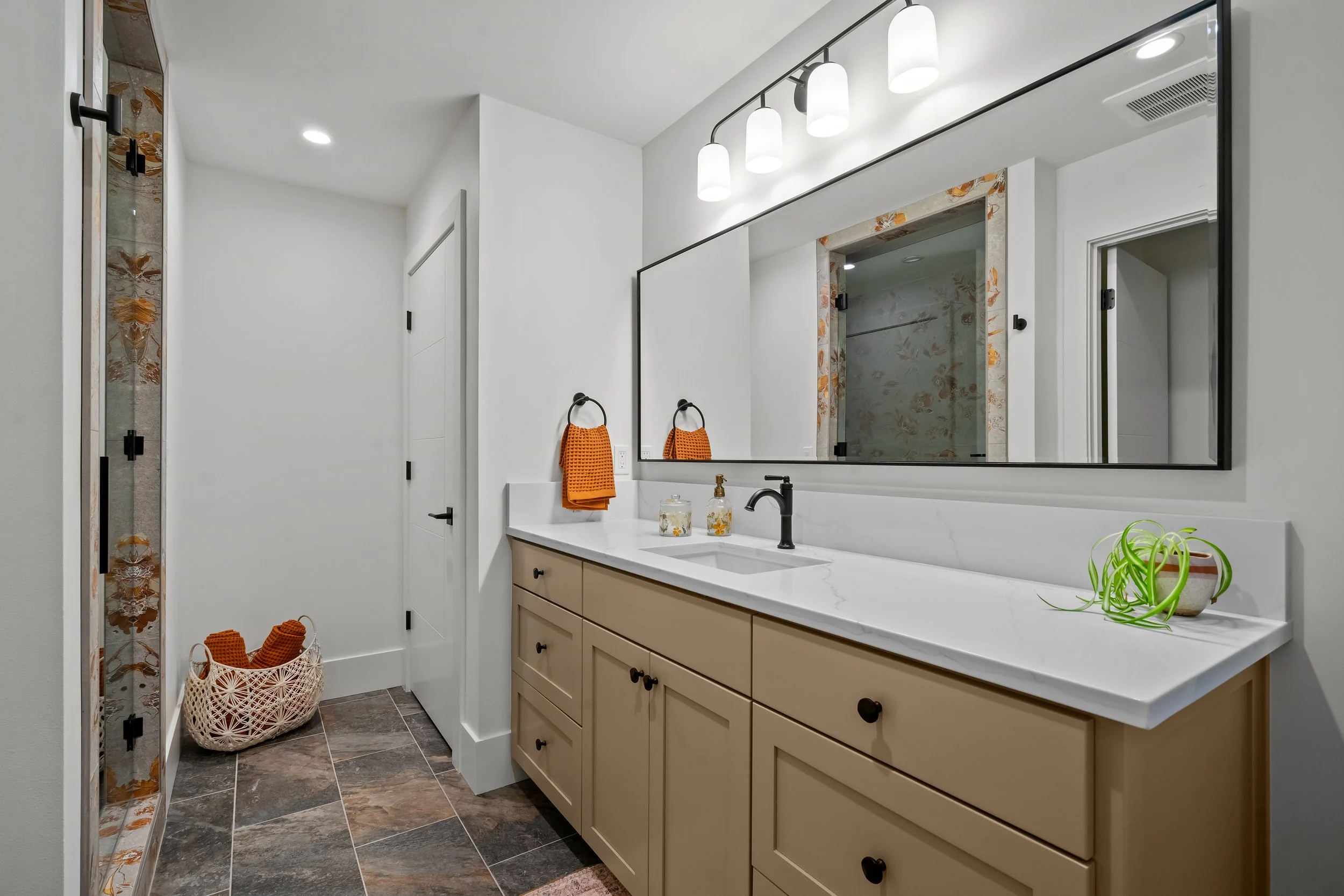 Modern bathroom with a large mirror, white marble countertop, beige cabinets, black fixtures, and a glass shower with floral accents. Orange towels and a decorative basket are on the floor, and a plant is on the countertop.