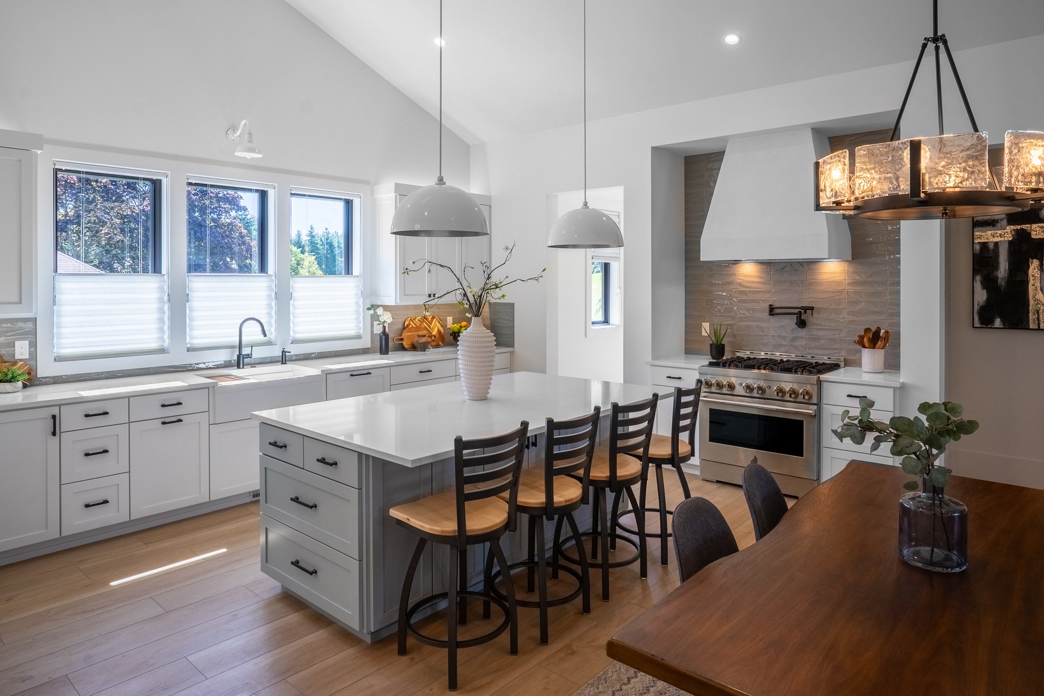 Modern kitchen with white cabinets, a kitchen island with a white surface, and a dining area with a wooden table and black chairs. There are three windows with white blinds, pendant lights hanging from the ceiling, and a stainless steel stove with a 