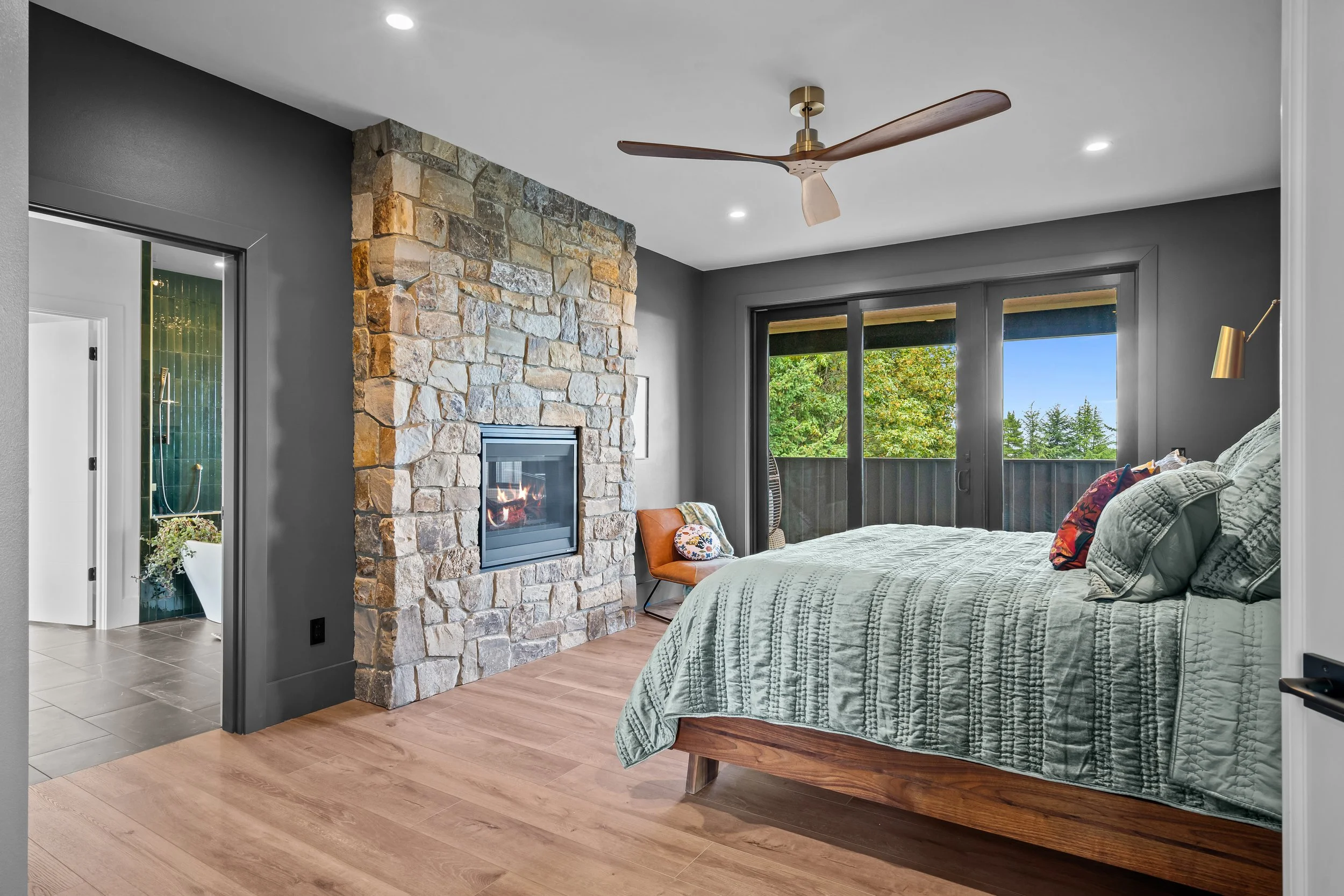 A bedroom with a stone fireplace, large windows showing greenery outside, a bed with a light green quilt, a wooden chair with a pillow, a wall-mounted lamp, and a ceiling fan.