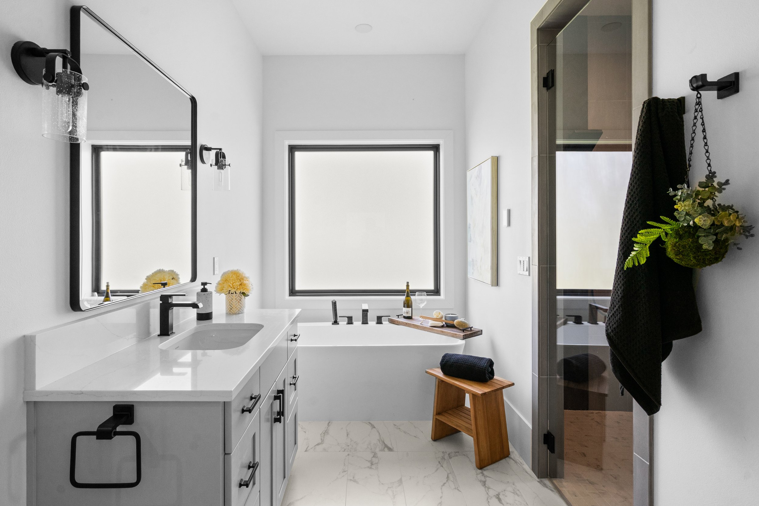 Modern bathroom with light blue cabinetry, black fixtures, a large mirror, a window above a soaking tub.