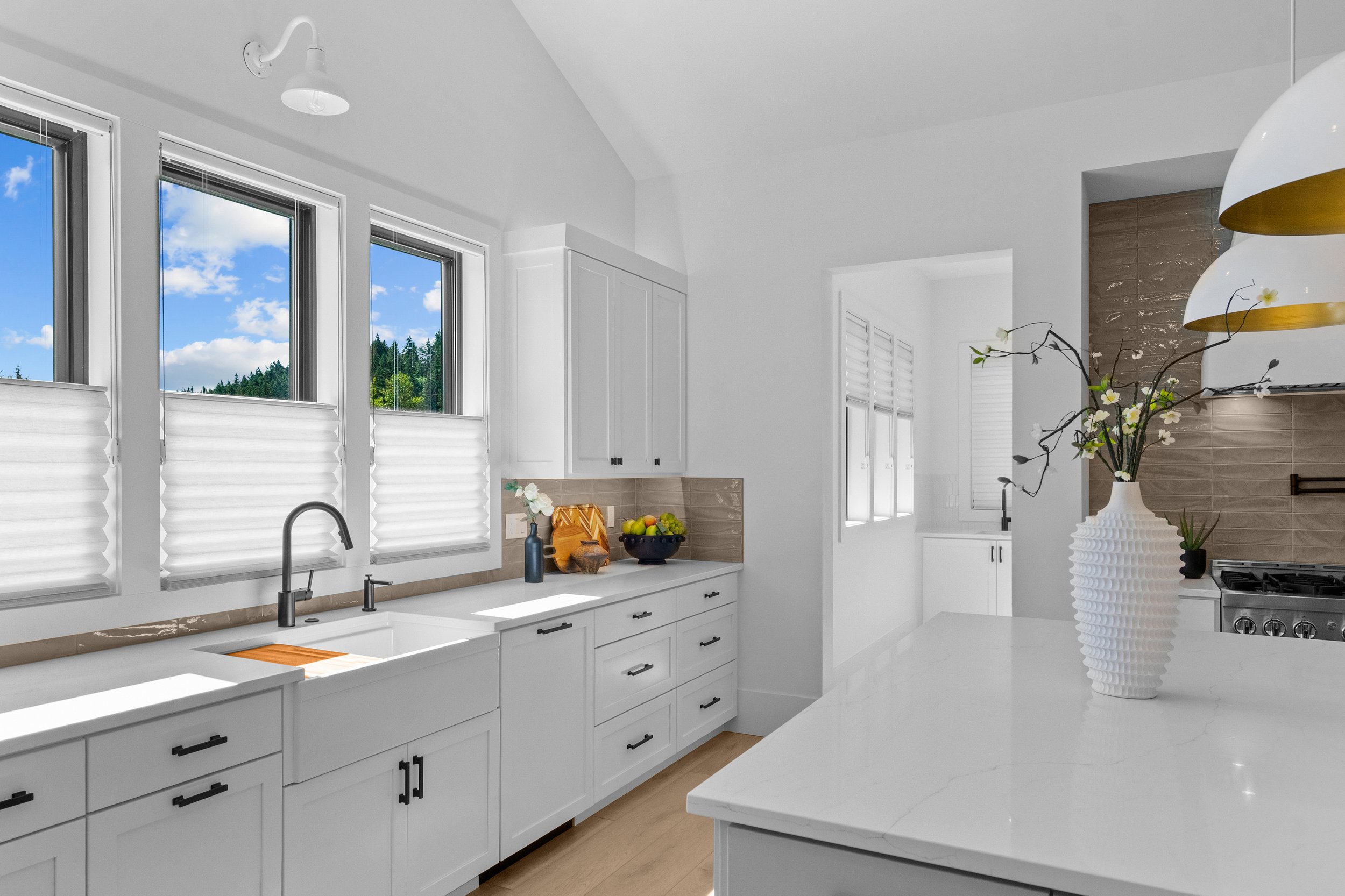 Modern farmhouse kitchen with white cabinets, marble countertops, pendant lights and three windows showing blue sky and trees outside. 