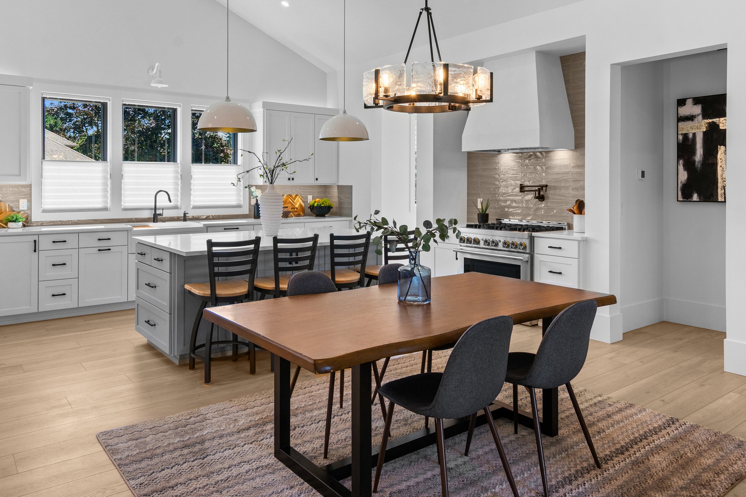 Modern kitchen with white cabinets, black hardware, and light wood flooring. Features a kitchen island with black chairs, a wooden dining table, and black chairs. Has large windows with white blinds, and pendant lights hanging from the ceiling. 