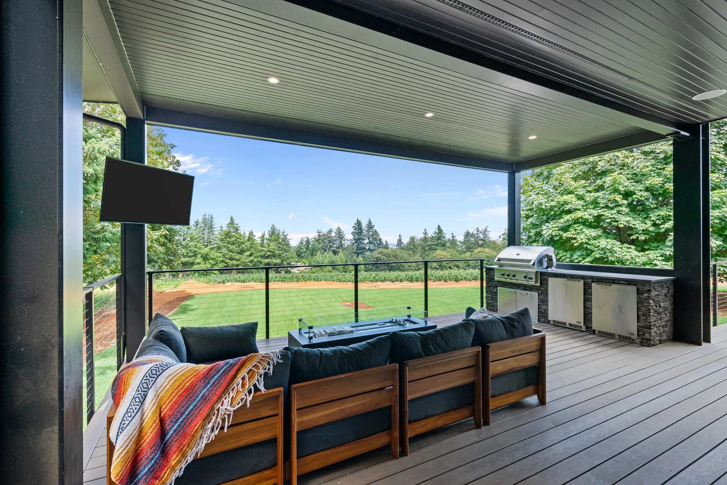 Outdoor patio with a roof, a mounted TV, a barbecue grill, a couch with pillows, and a scenic view of trees and blue sky.
