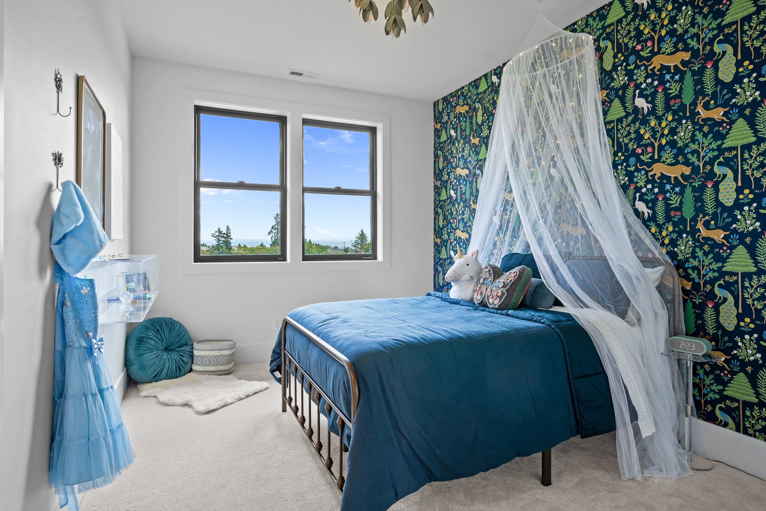 A bedroom with white walls and a large window showing trees and a blue sky outside. The bed has a blue comforter and a stuffed horse toy, with a white mosquito net over it. There's a black digital clock on a nightstand displaying '2:23', a small fram