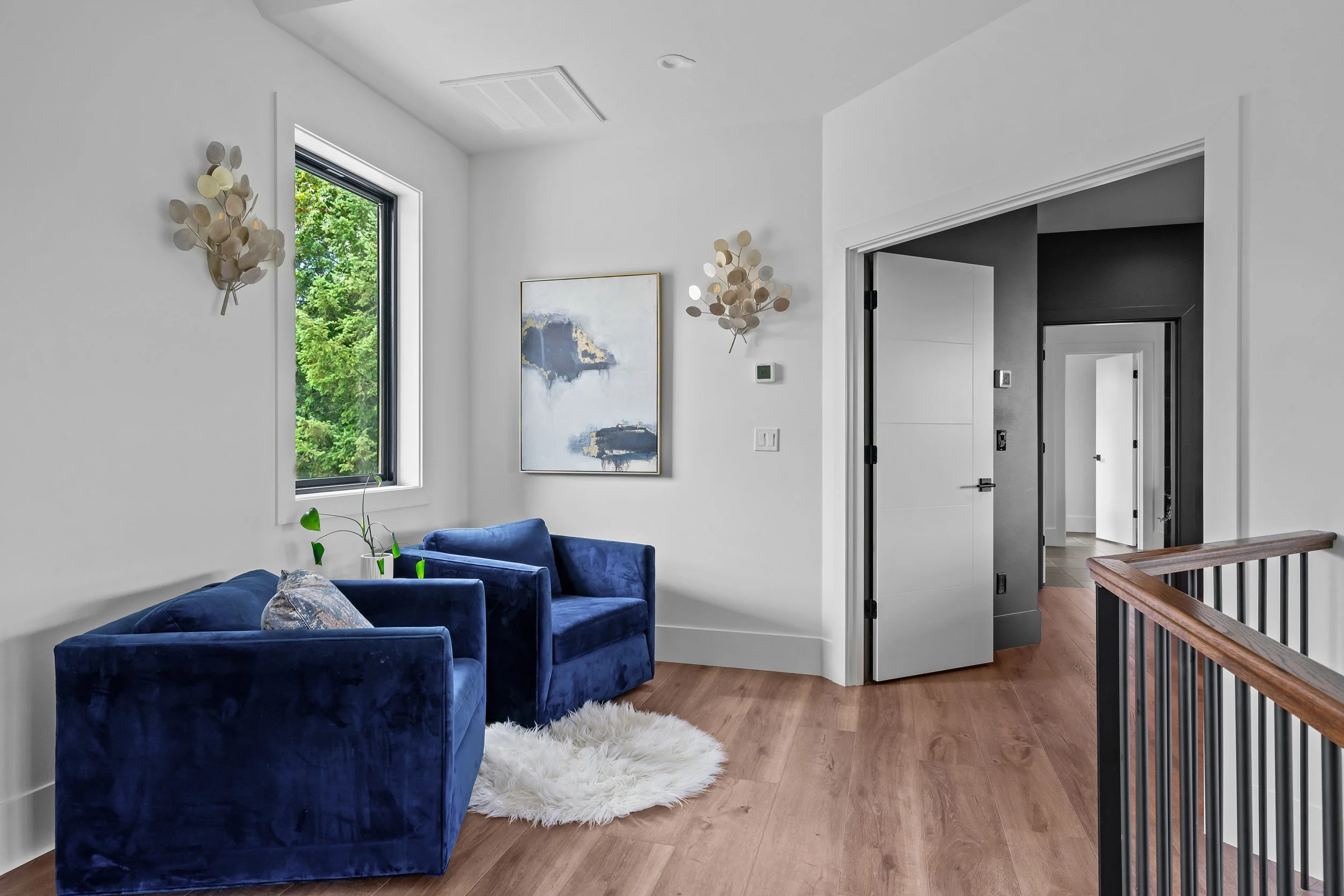 Sitting area with white walls, a large window showing green trees outside, two navy blue velvet sofas, a white fluffy rug, wall art, wall-mounted light fixtures, wooden flooring, and a black-and-white doorway leading to other rooms.